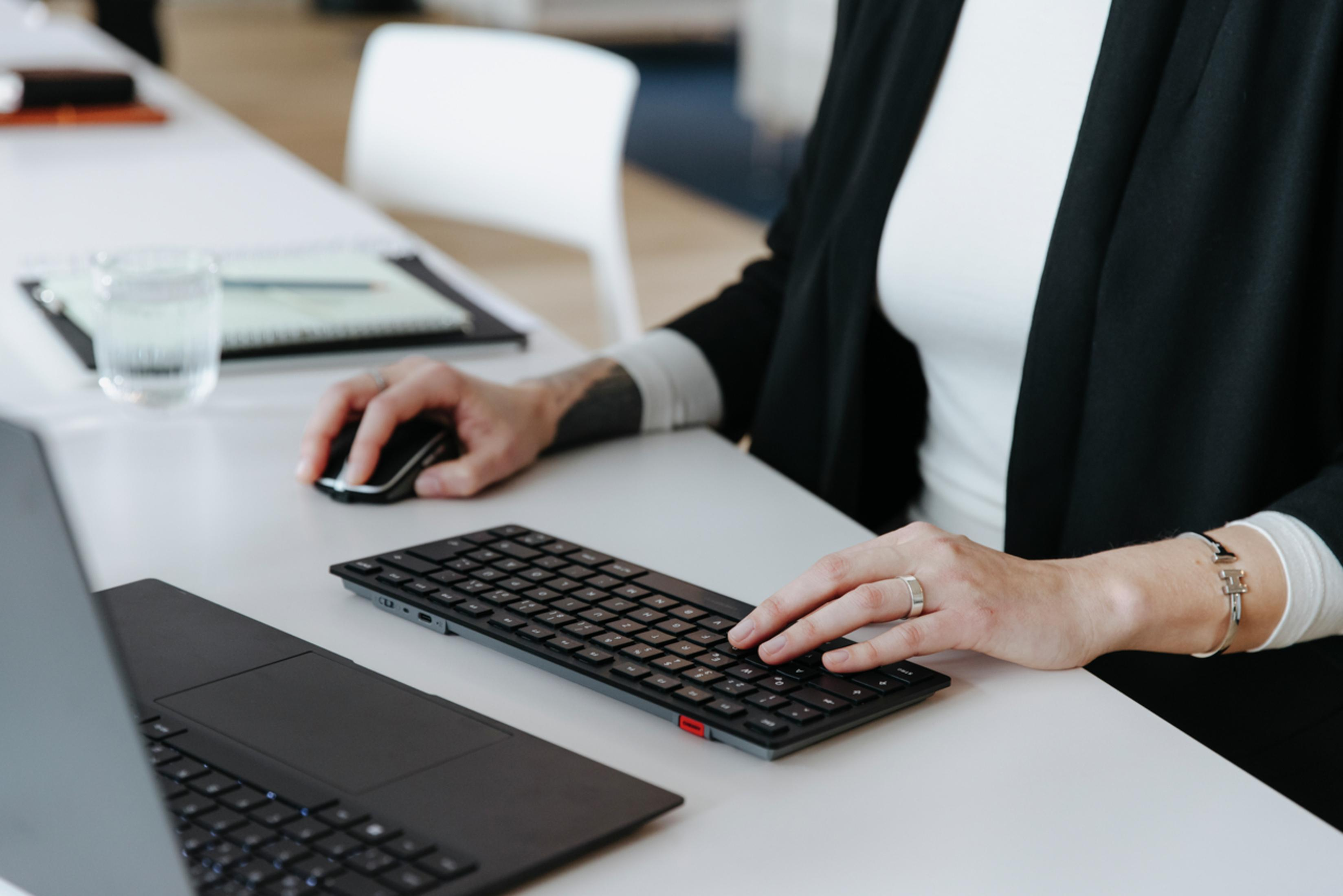 Osoba korzystająca z komputera, pisząca na klawiaturze i używająca myszy, na białym biurku.