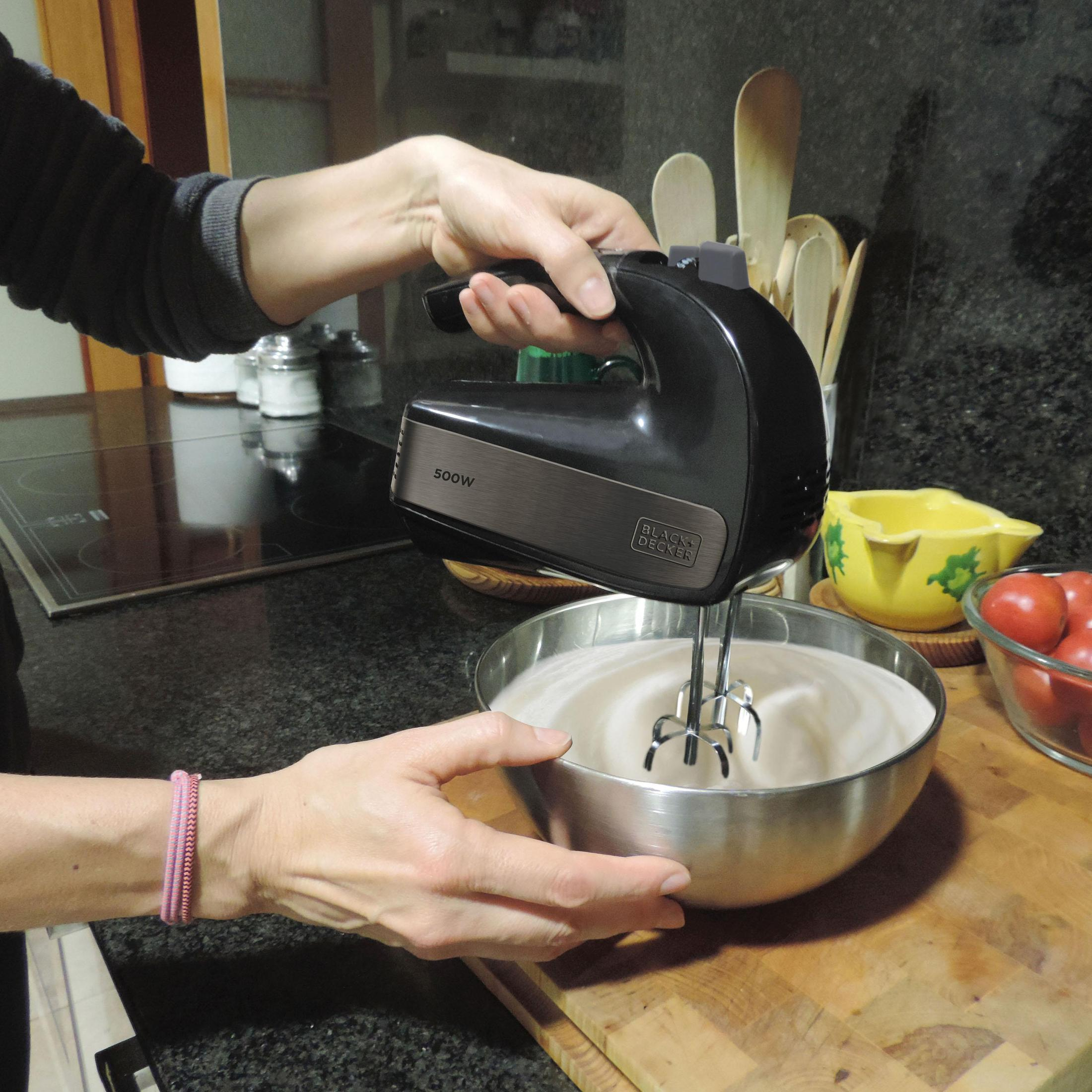 Eine Person benutzt einen schwarz-kupferfarbenen Handmixer, um etwas in einer Metallschüssel zu schlagen.
