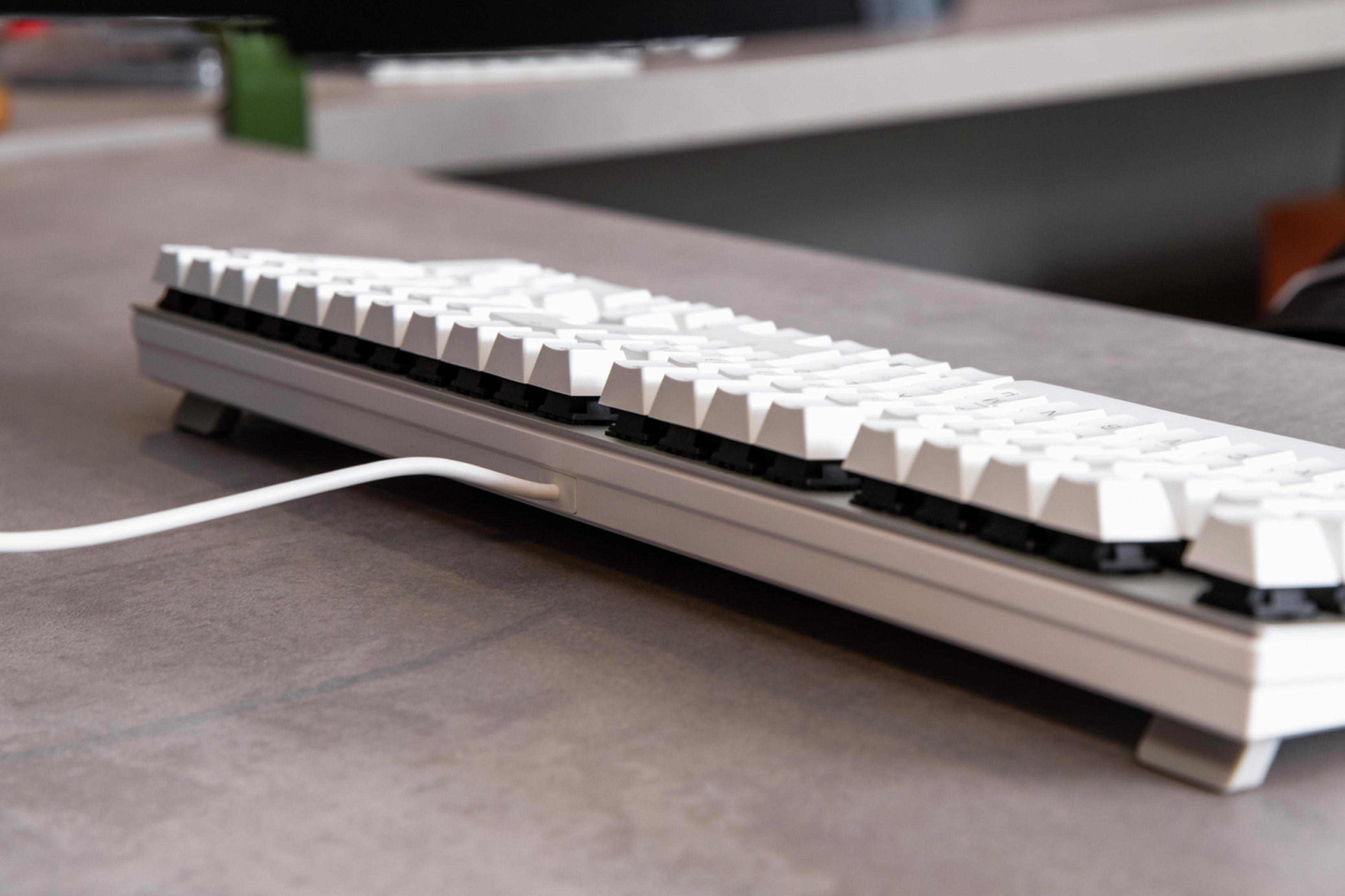 Eine weiße Tastatur mit schwarzen Tasten auf einem grauen Schreibtisch, ein weißes Kabel ist sichtbar.