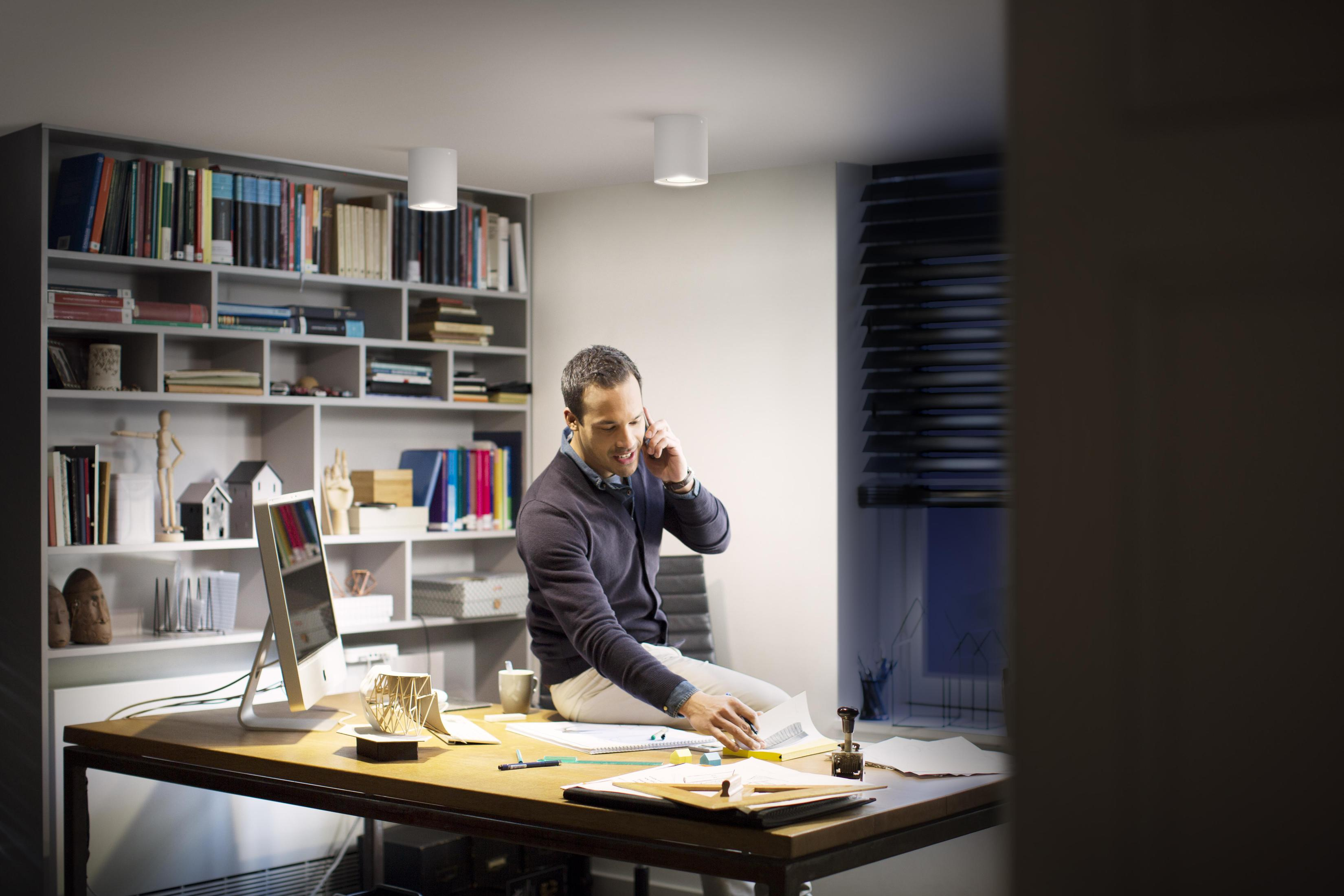 Un homme assis sur un bureau, parlant au téléphone, entouré de papiers et d'un ordinateur.