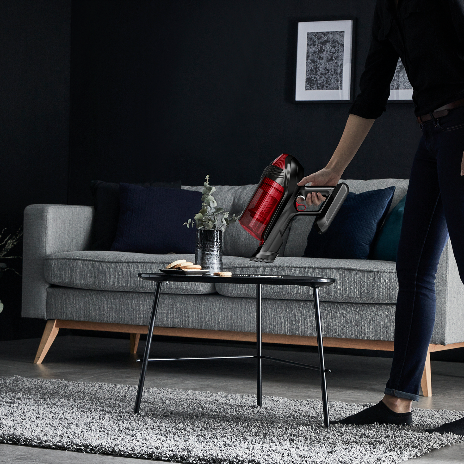 Person saugt einen Teppich mit einem kabellosen Staubsauger in Schwarz und Rot, in der Nähe eines Tisches und Sofas.