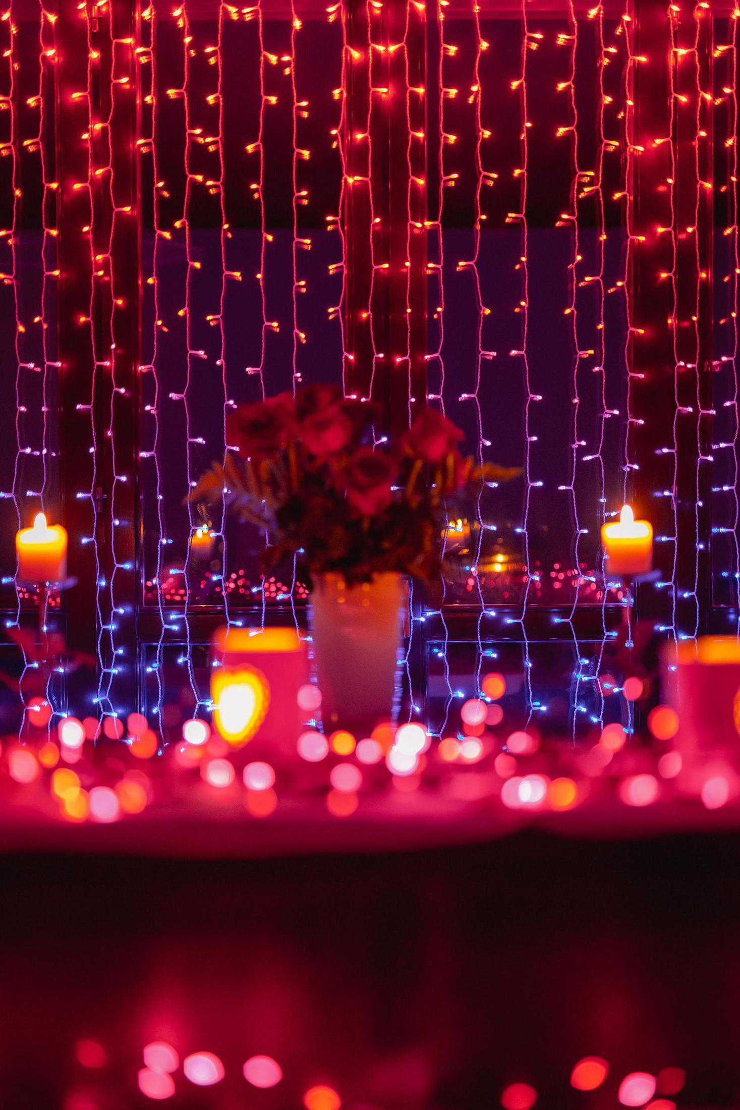 Okno z czerwonymi i niebieskimi lampkami choinkowymi, świecami i wazonem kwiatów.