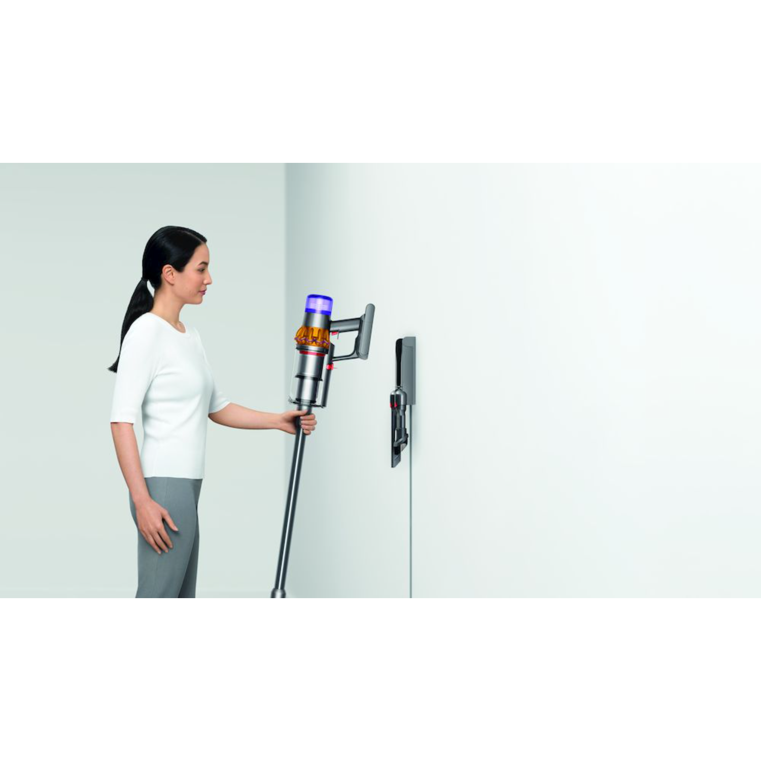 Kobieta w białej koszuli używa srebrnego odkurzacza Dyson. Na ścianie znajduje się stacja ładująca.