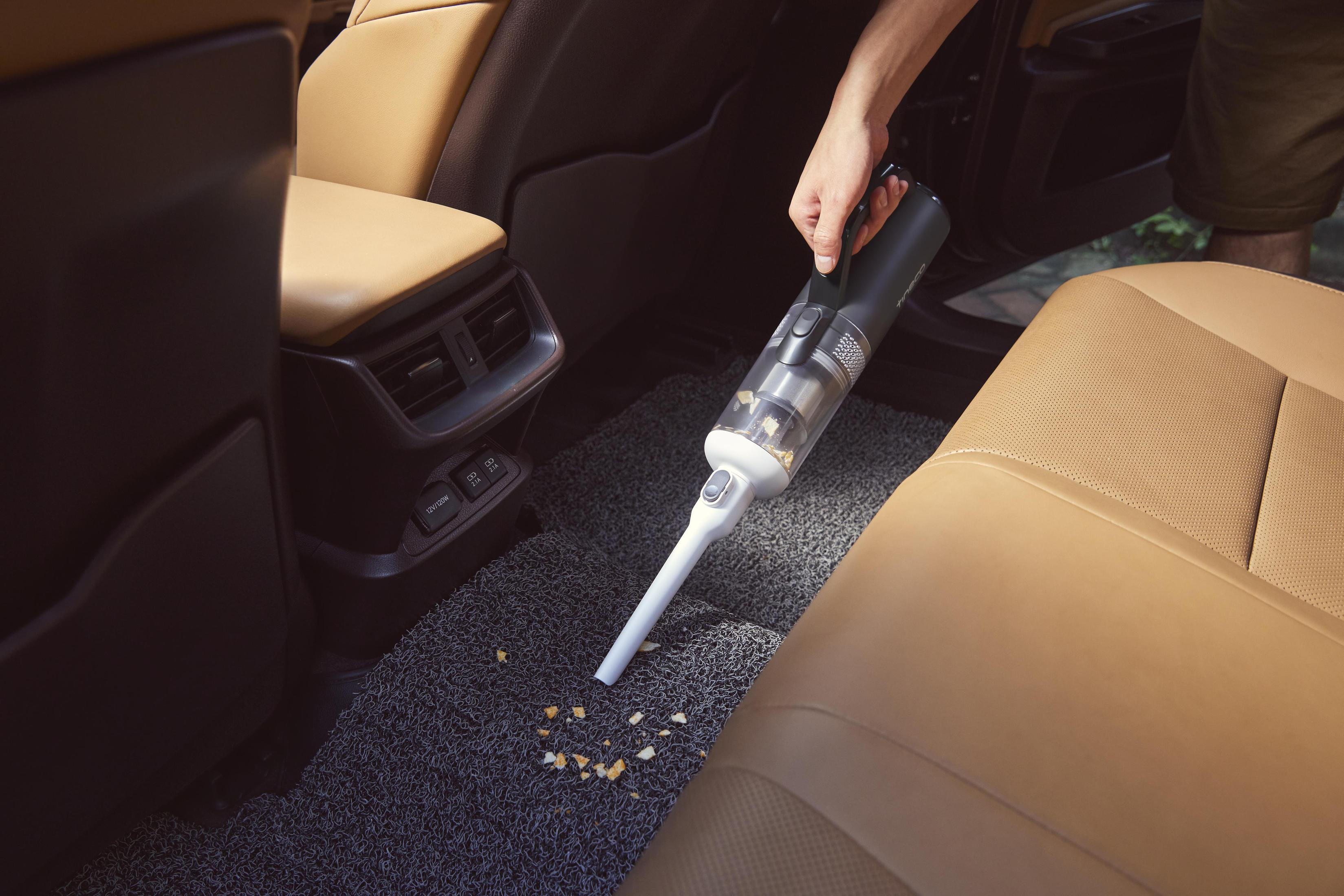 Ein Handstaubsauger, der einen Autoinnenraum saugt; Krümel auf dem Boden, beige Sitze und dunkler Innenraum.