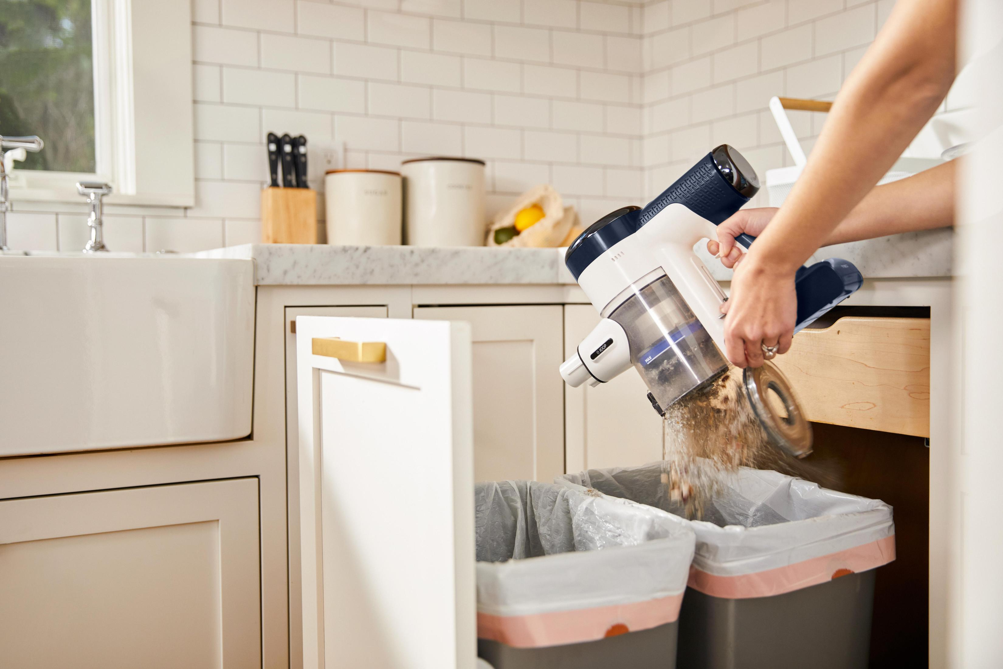 Una persona vacía una aspiradora inalámbrica en dos botes de basura en una cocina blanca.