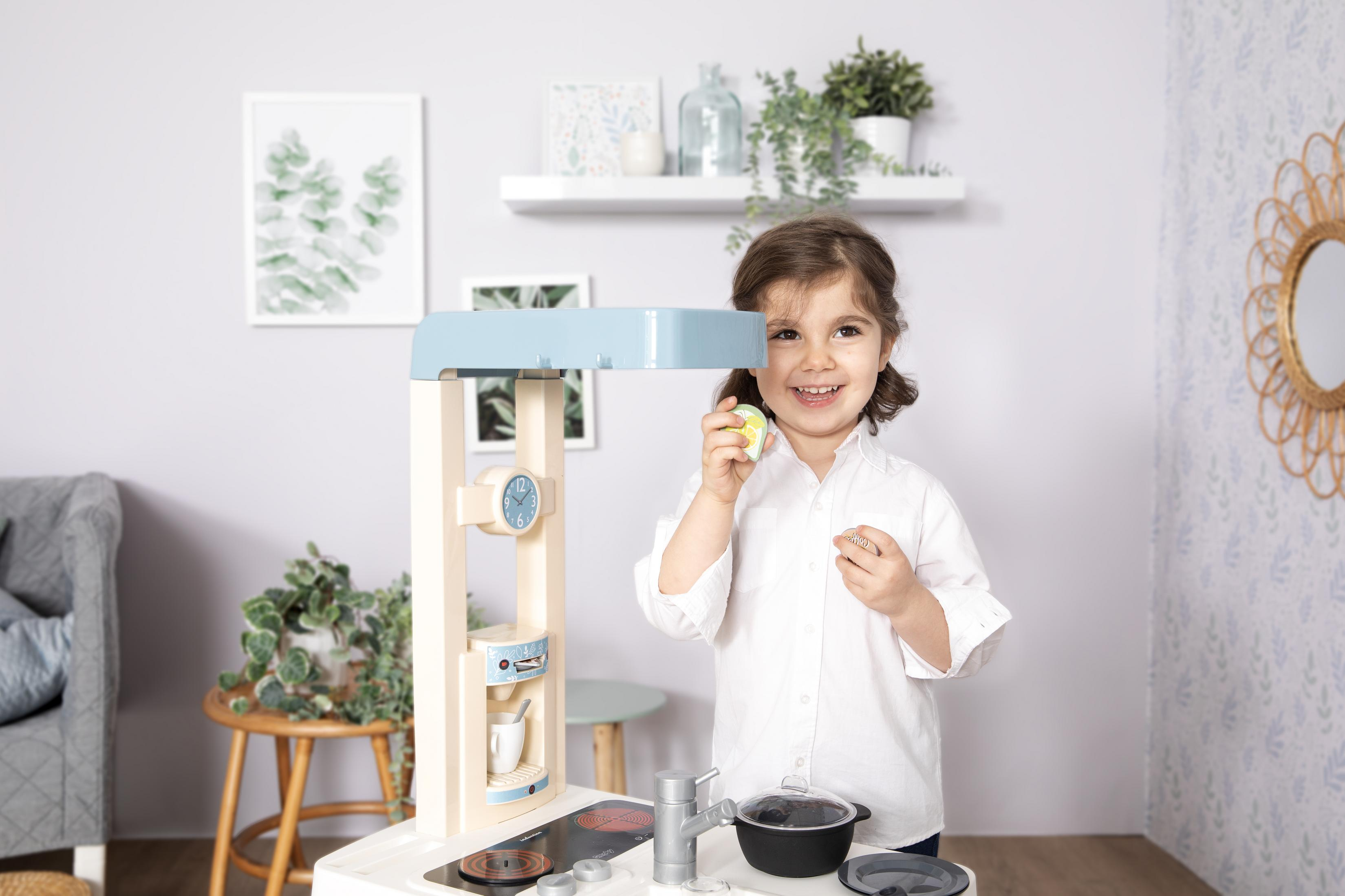 Mała dziewczynka bawi się zestawem kuchennym w jasno oświetlonym pokoju. Uśmiecha się.
