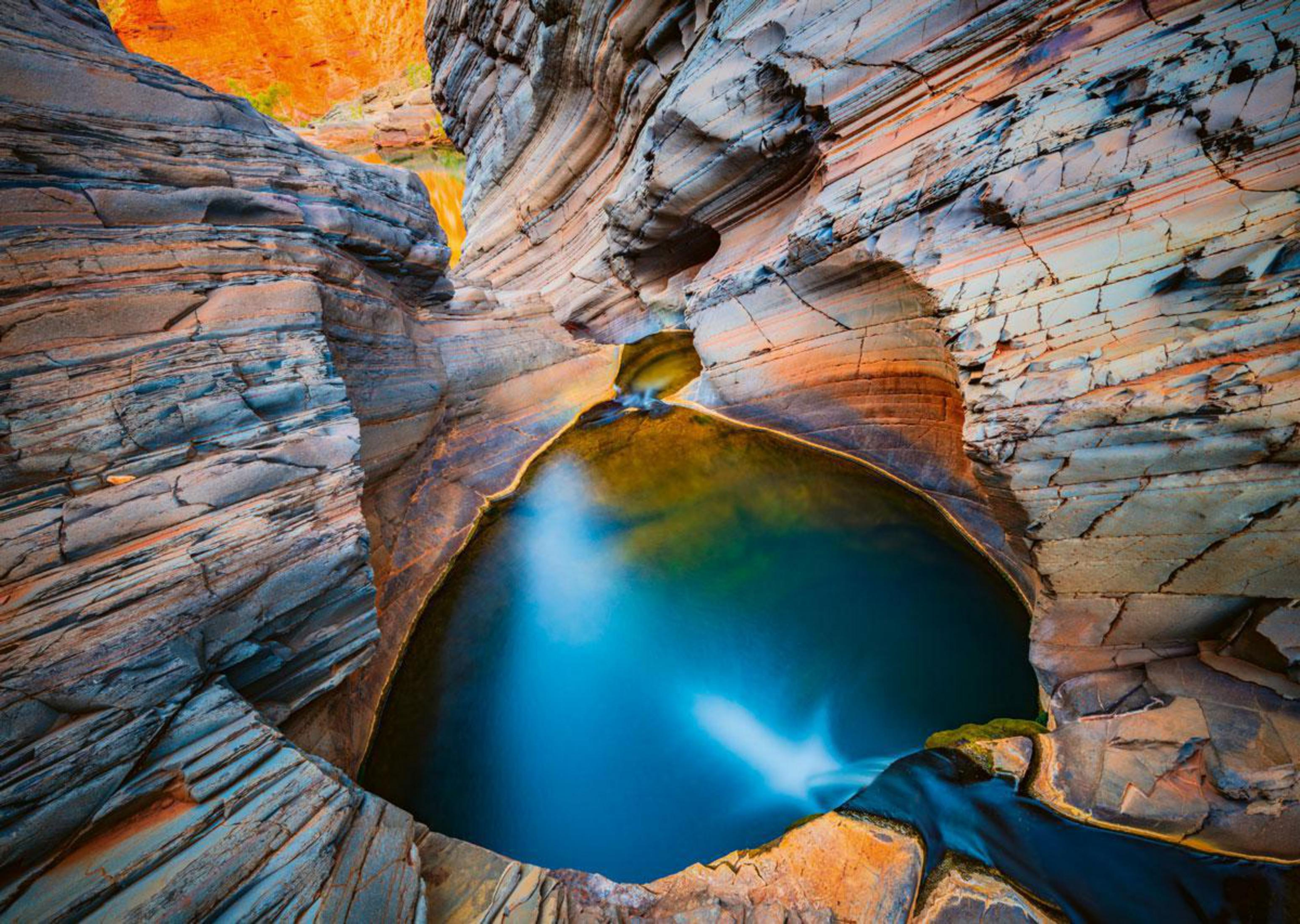 Un cañón con formaciones rocosas en capas y una piscina de agua azul en el centro.