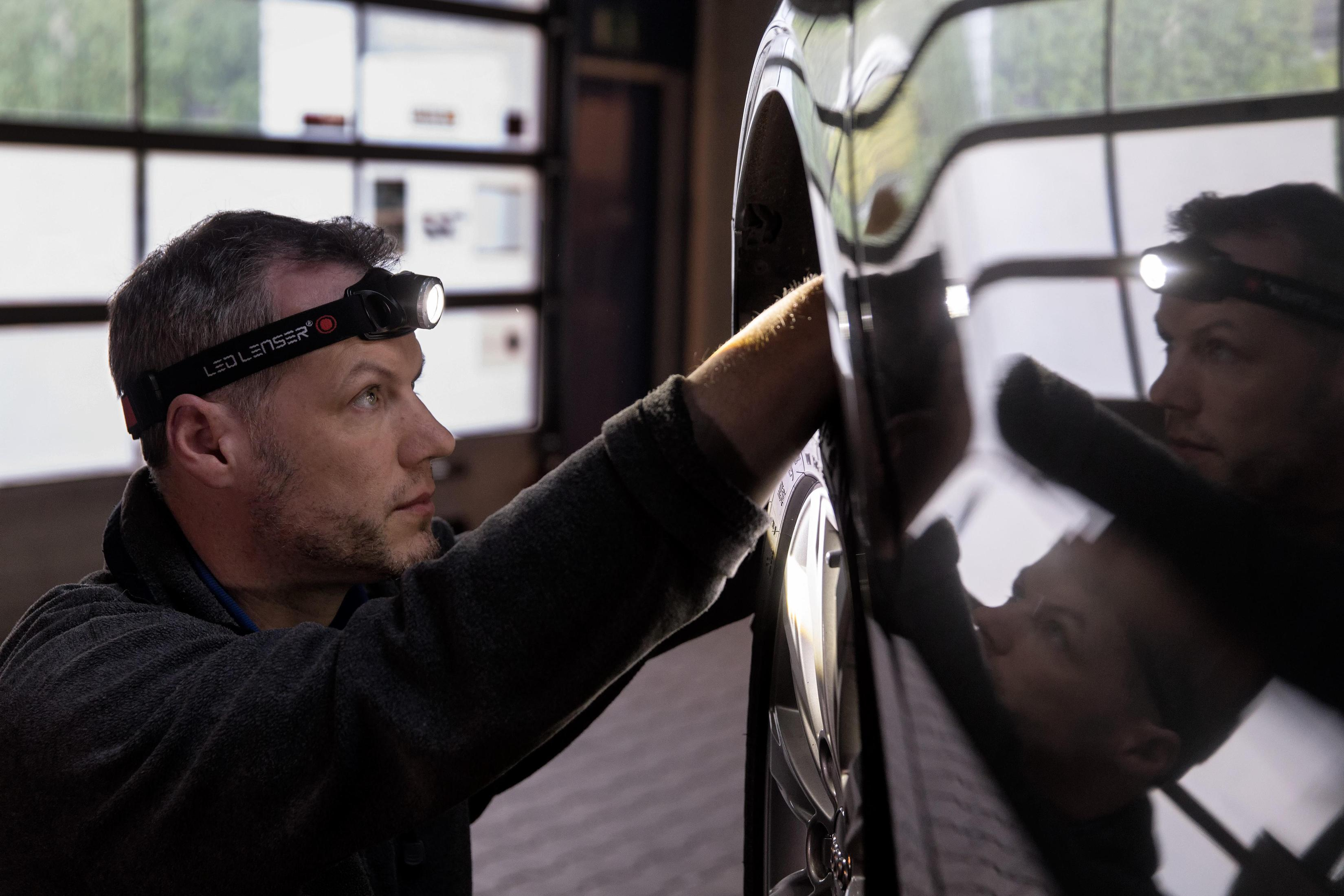 Mężczyzna w lampie czołowej sprawdza oponę samochodową, widoczny jego odbicie.