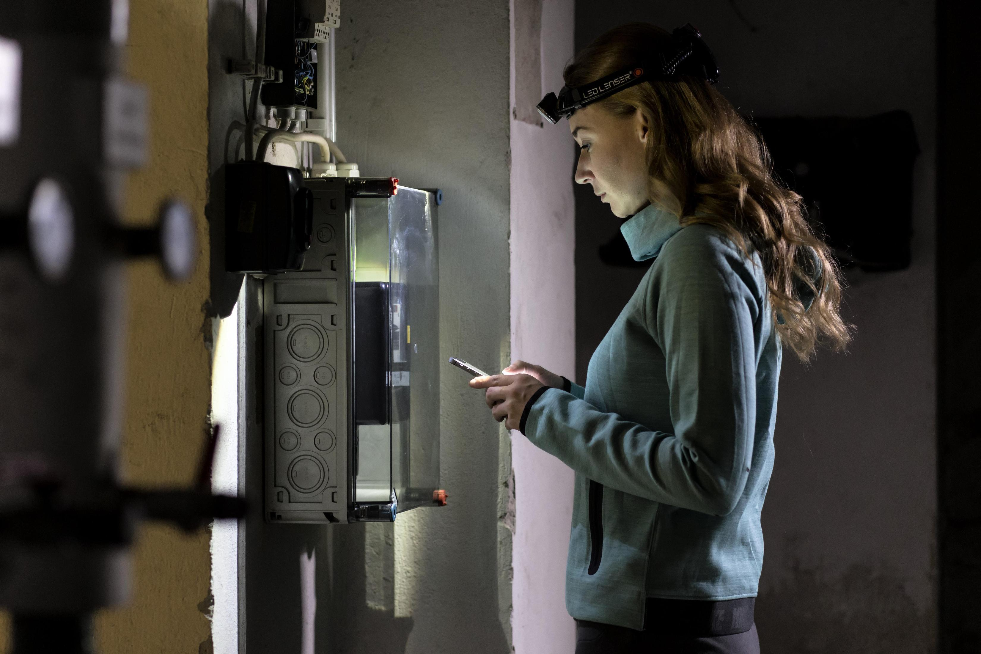 Kobieta z czołówką używa telefonu obok panelu elektrycznego.