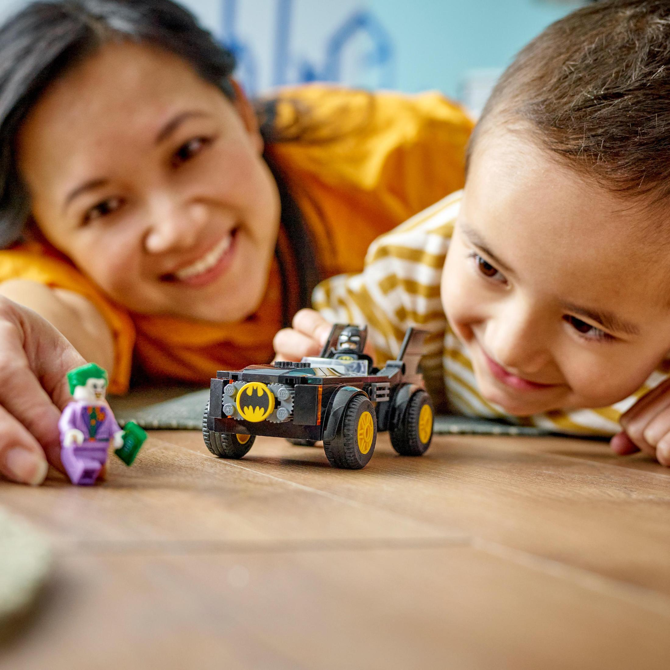 Eine Mutter und ein Kind spielen mit Lego Batman-Auto und Joker-Figur auf einem Holzboden.
