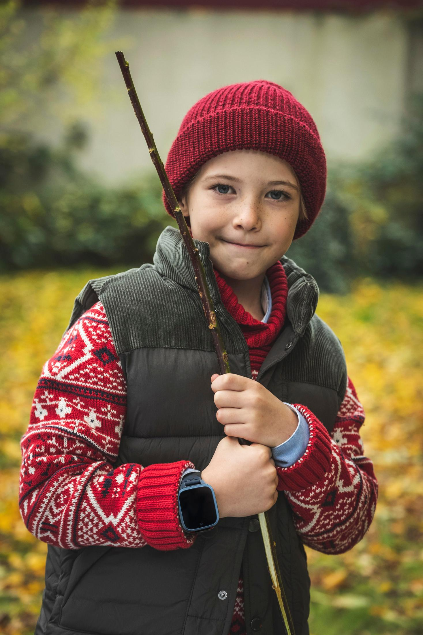 Junge mit roter Mütze und Pullover, hält Stock, lächelt im Herbst.