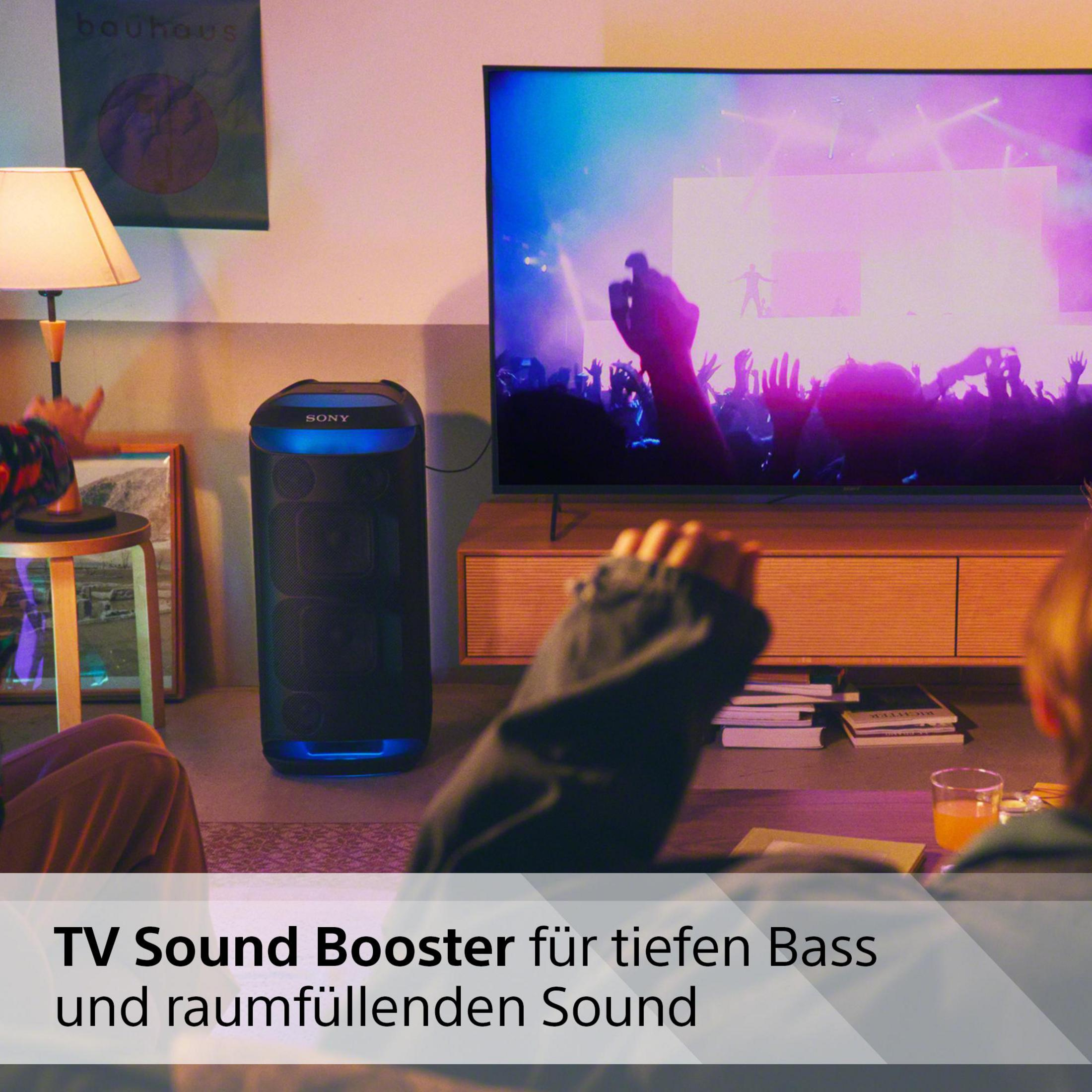 Ein Fernseher zeigt ein farbenfrohes Konzert. Ein schwarzer Lautsprecher darunter. Menschen schauen zu.