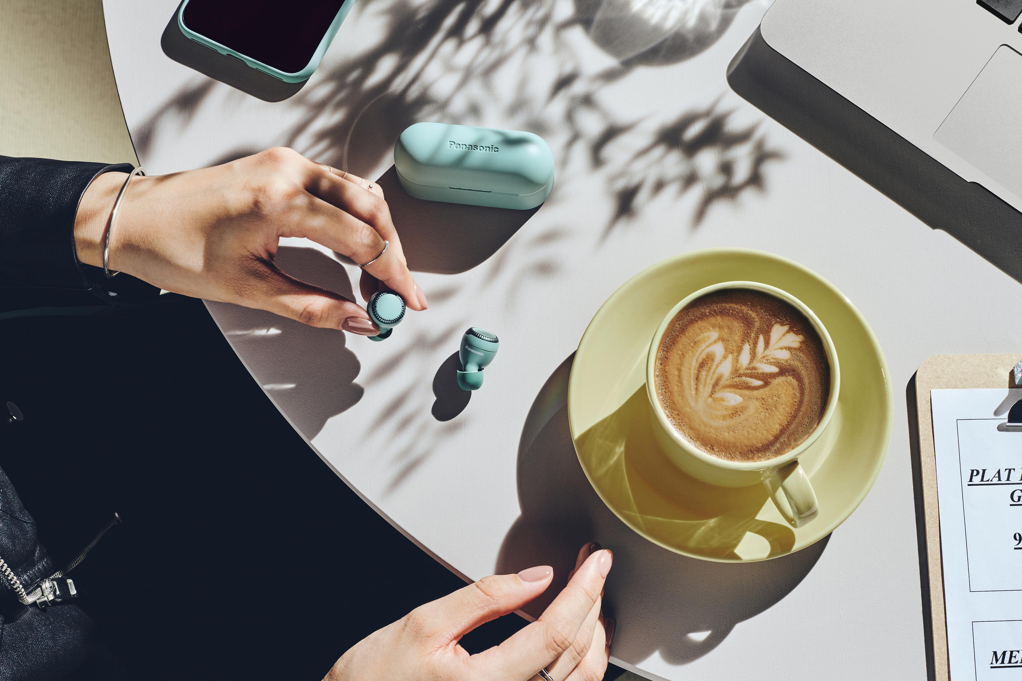 Hände halten Ohrhörer und Etui auf einem Tisch mit einer Kaffeetasse und einem Laptop.