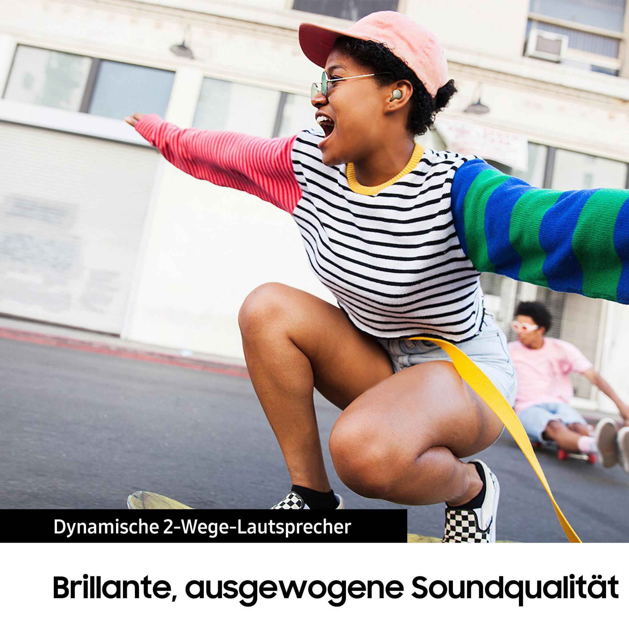 Eine Frau auf einem Skateboard mit Ohrhörern, lächelnd, und eine andere Person im Hintergrund.