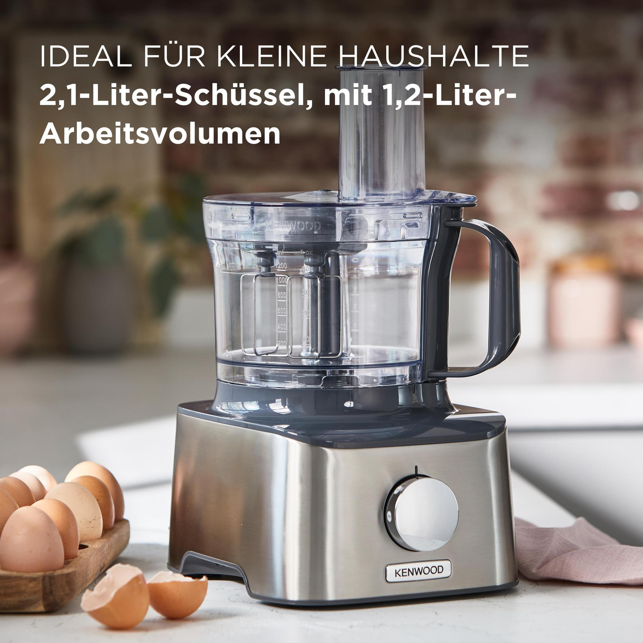 Silber- und grauer Küchenroboter mit transparentem Behälter, neben Eiern und Eierschalen auf einem Tisch.