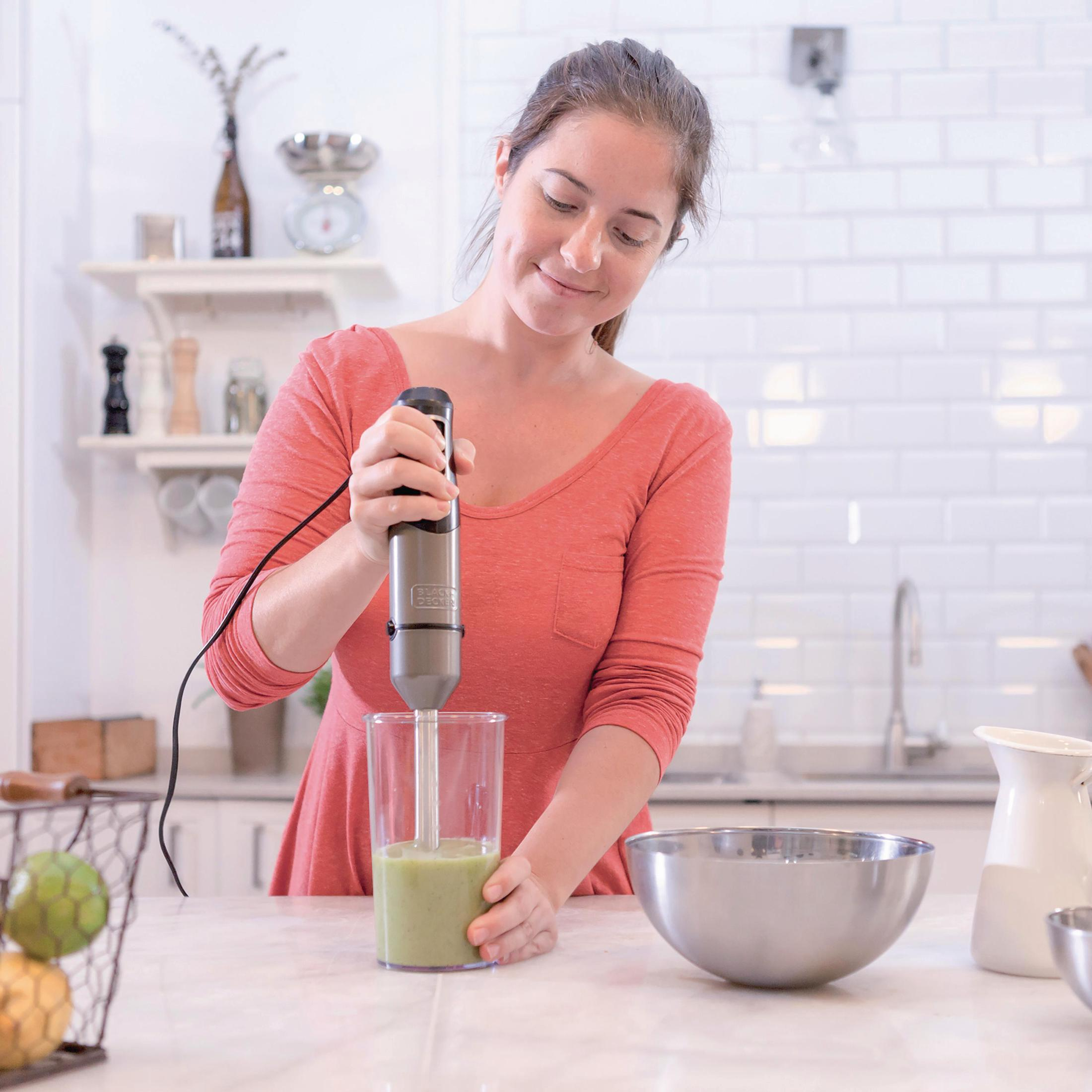 Kobieta używa szarego blendera ręcznego do mieszania zielonej substancji w szklance.