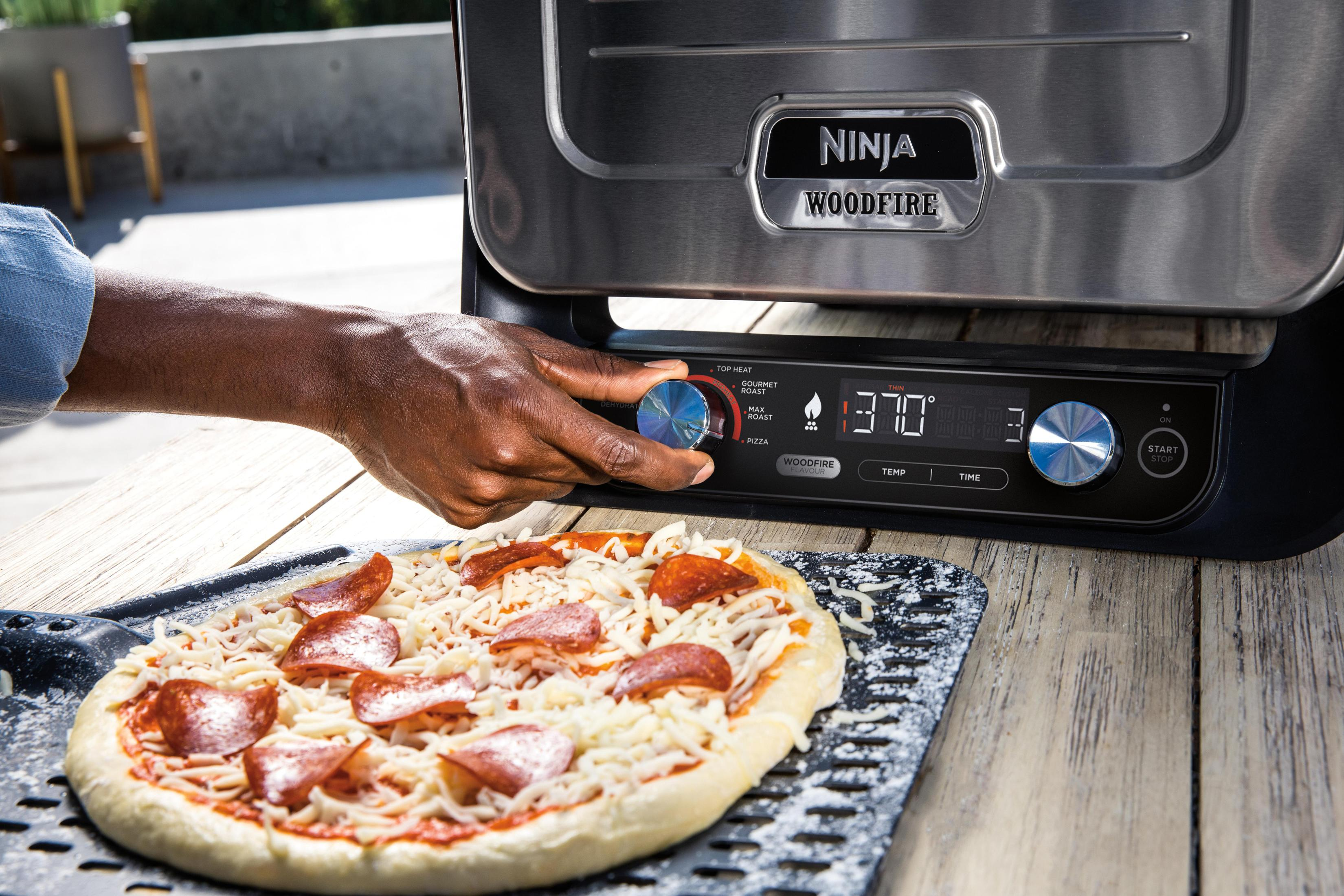 Une personne règle un cadran sur un grill Ninja Woodfire, une pizza est prête. Le grill est sur une terrasse en bois.