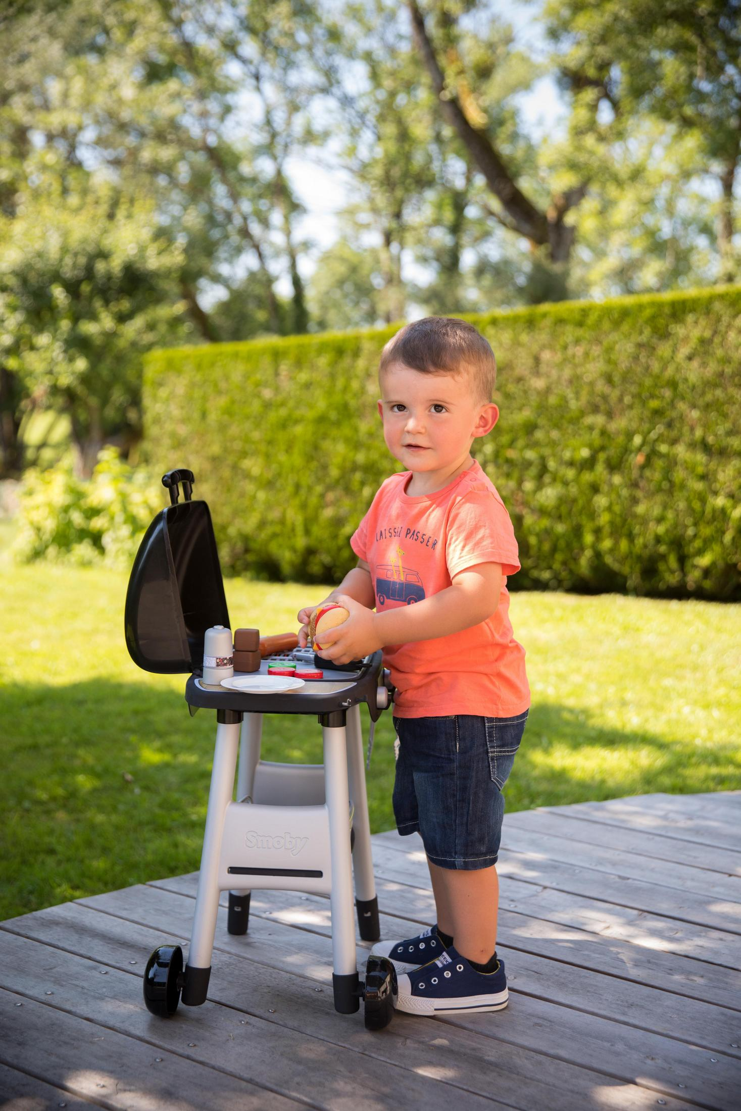 Ein kleiner Junge spielt mit einem Spielzeuggrill. Er hält eine Spielzeugwurst und trägt ein T-Shirt.