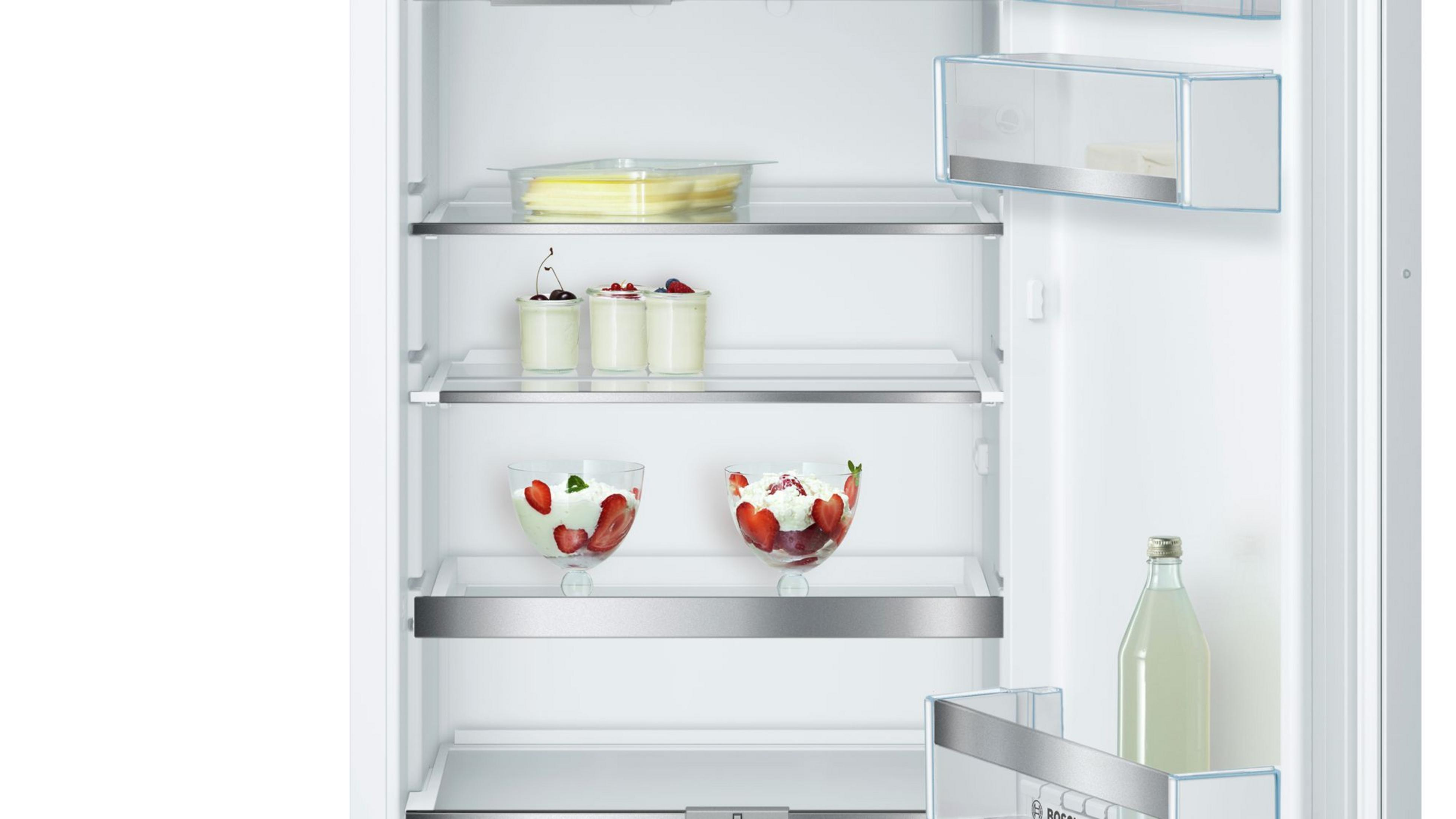 Offener Kühlschrank mit Regalen, die mit Lebensmitteln gefüllt sind: Desserts, Früchte und eine Wasserflasche.