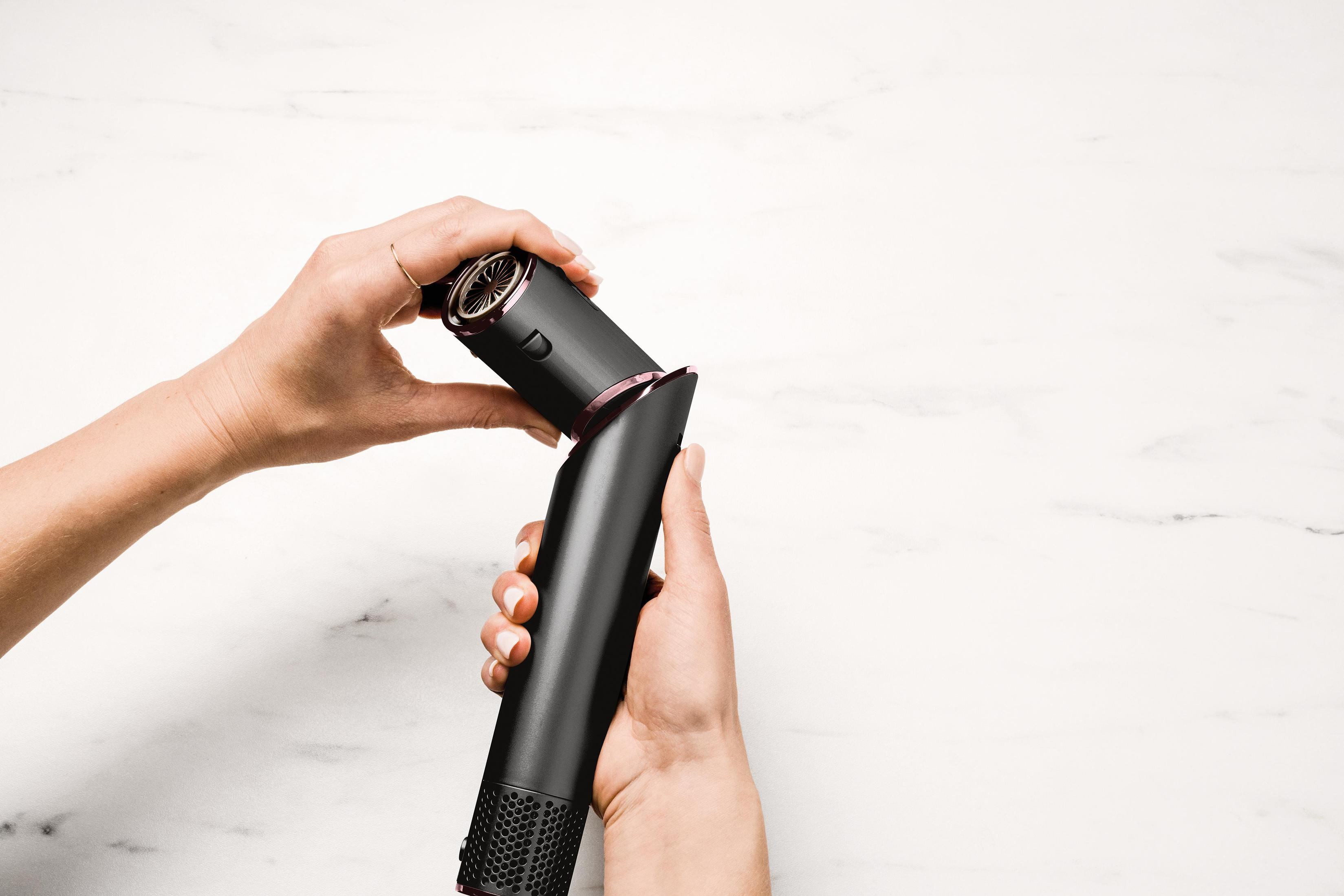 Dłonie kobiety trzymają czarną prostownicę do włosów, rozdzielając ją. Białe marmurowe tło.