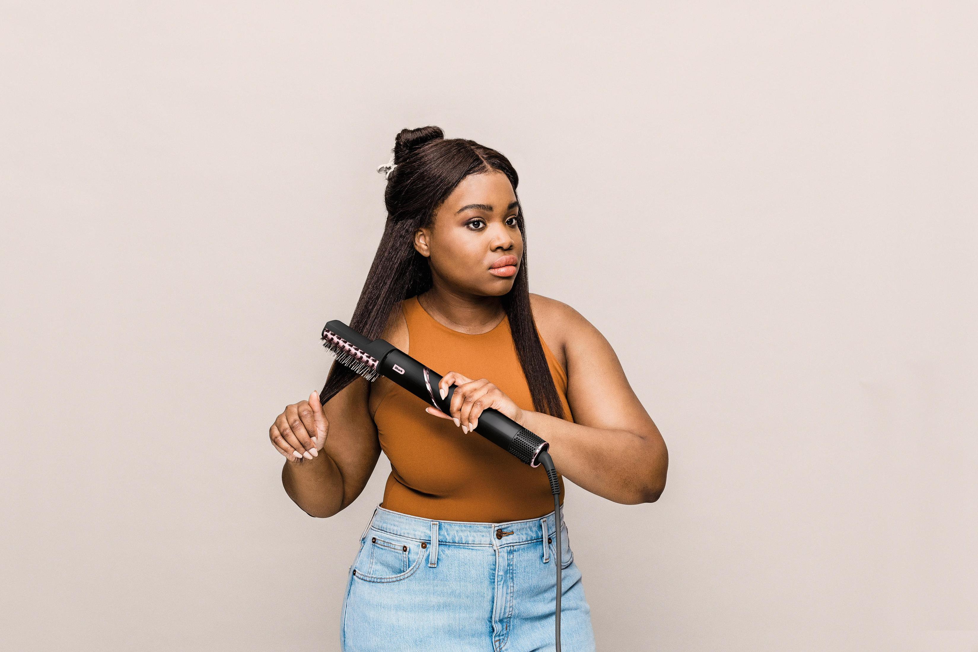 Kobieta z ciemnymi włosami używa czarnej szczotki do stylizacji. Beżowe tło.