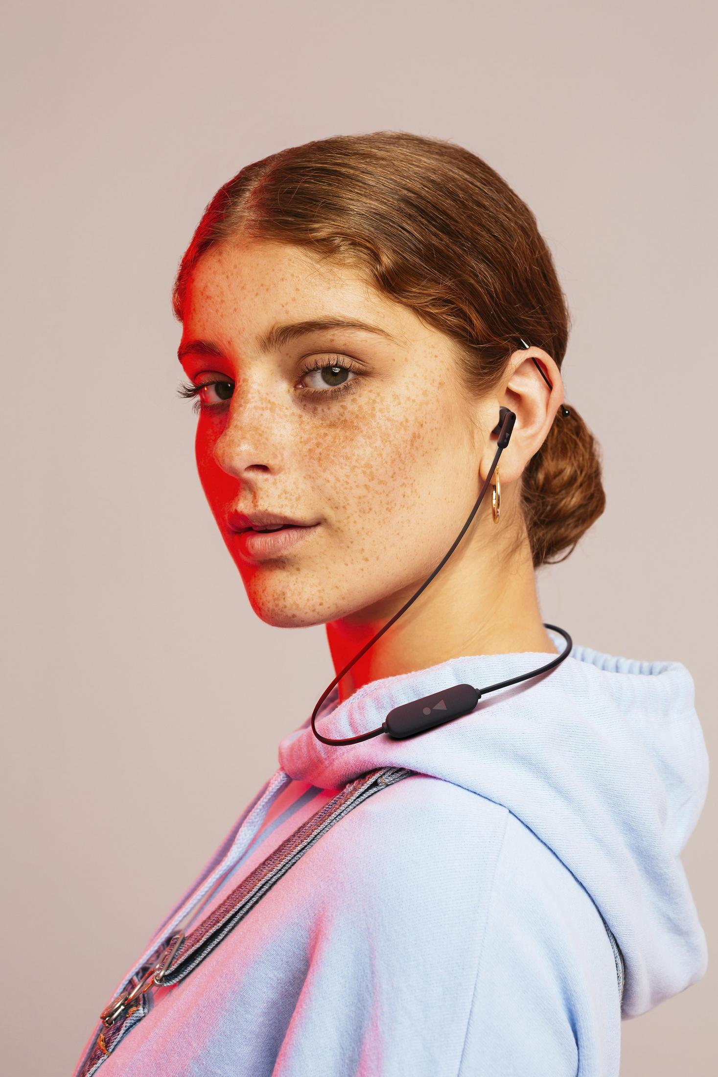 Młoda kobieta w niebieskiej bluzie z kapturem i słuchawkami, z czerwonym światłem na twarzy, patrzy w kamerę.