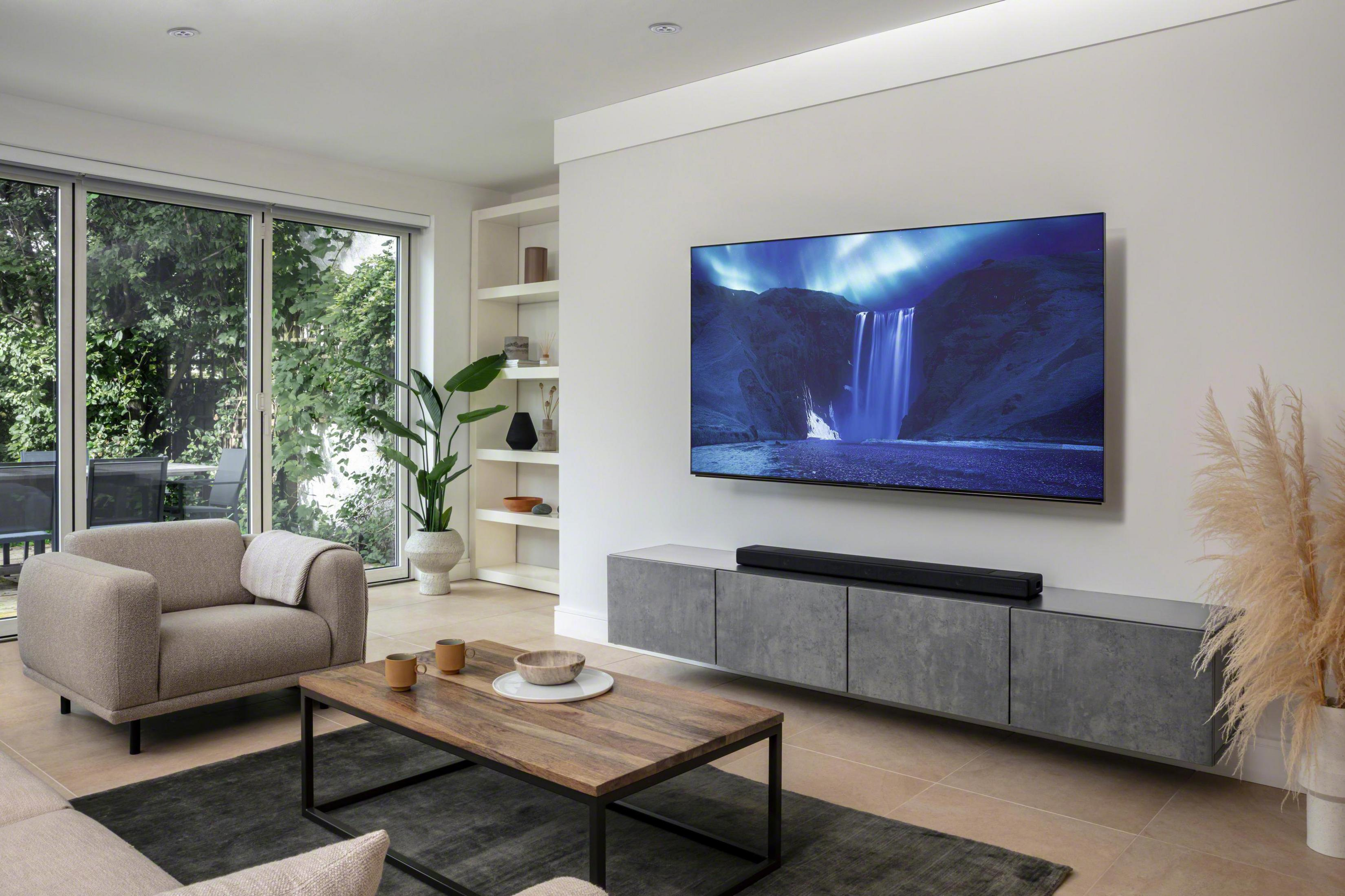 Wohnzimmer mit Fernseher an der Wand, Sofa und Couchtisch. Große Fenster und ein Teppich.