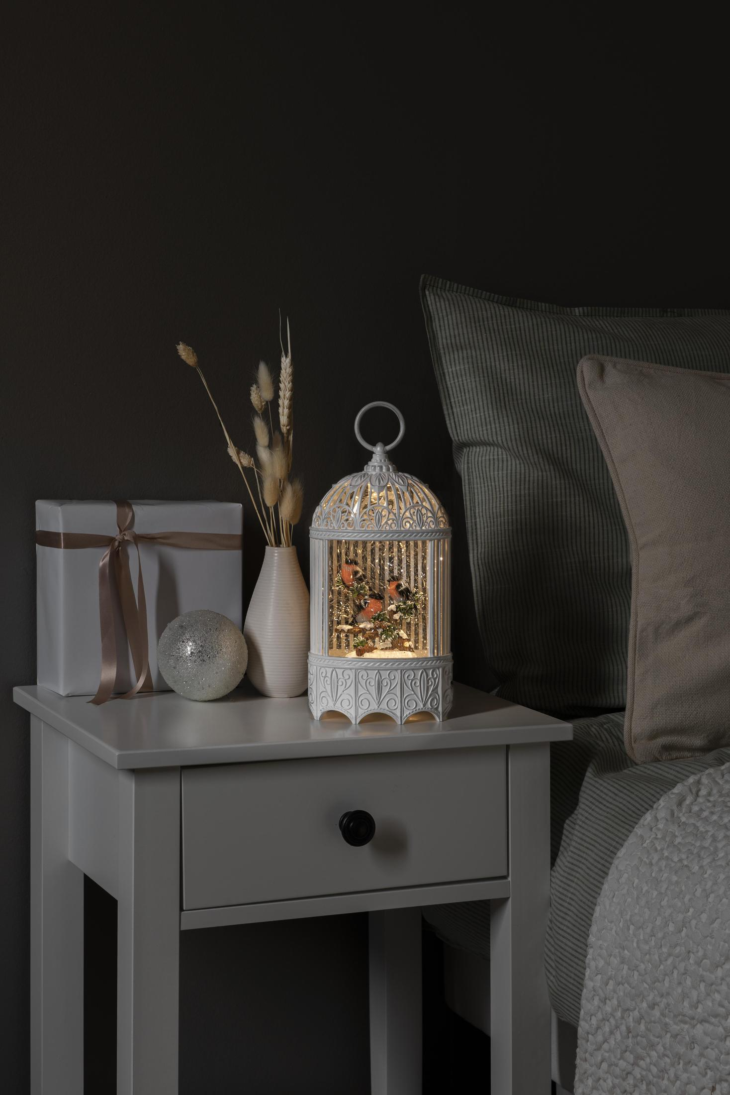 Ein weißer dekorativer Vogelkäfig mit einem Geschenk, einer Vase und einem Bett im Hintergrund.