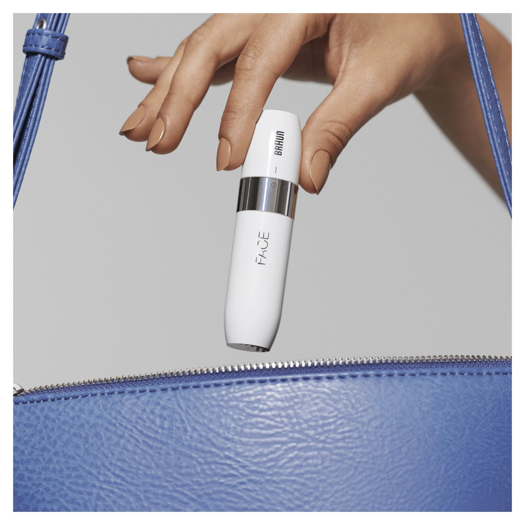 La mano di una donna tiene un dispositivo Braun Face bianco, vicino a una borsa blu con cerniera.
