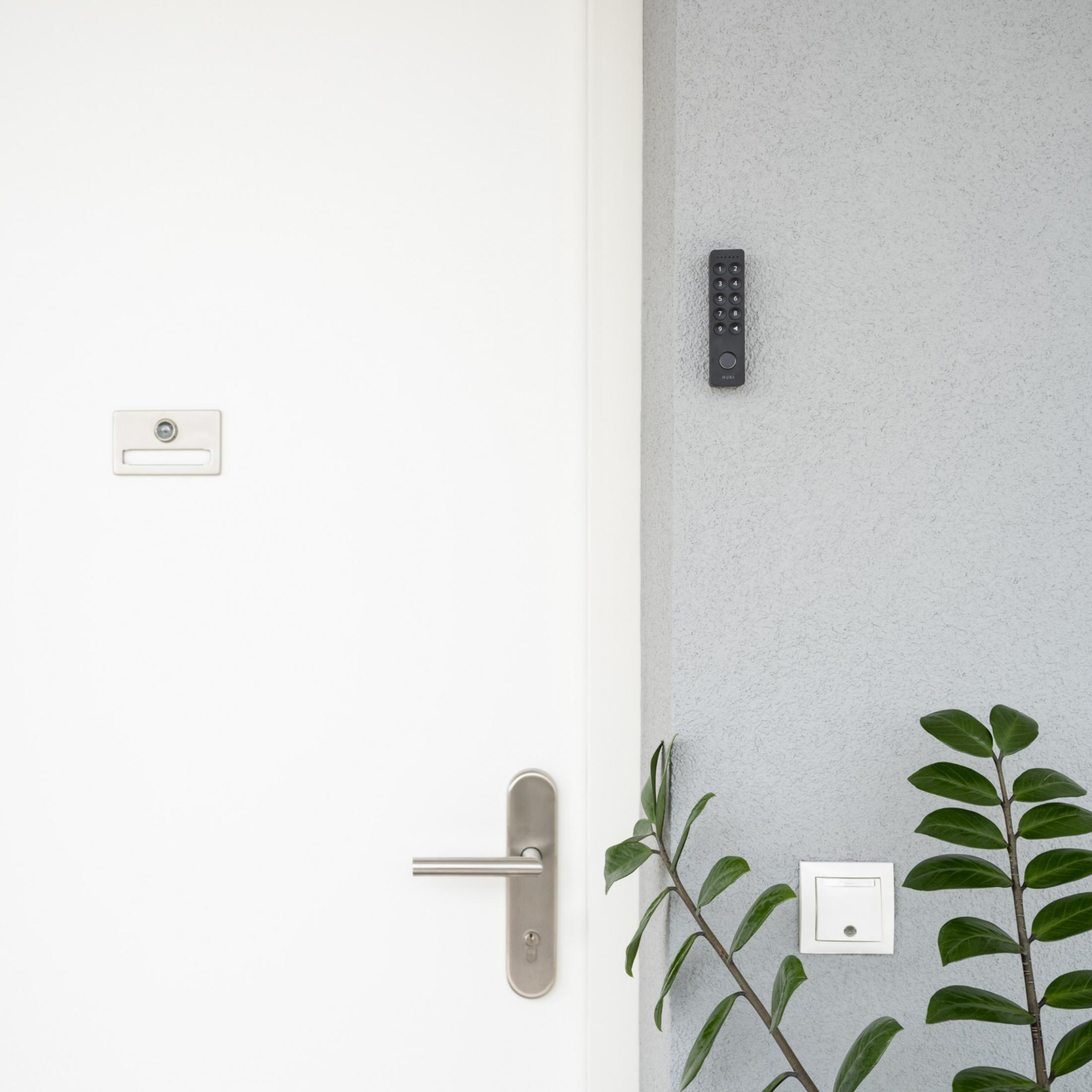 Een witte deur met een slot en een sleutelpaneel. Een afstandsbediening en een groene plant aan de muur.