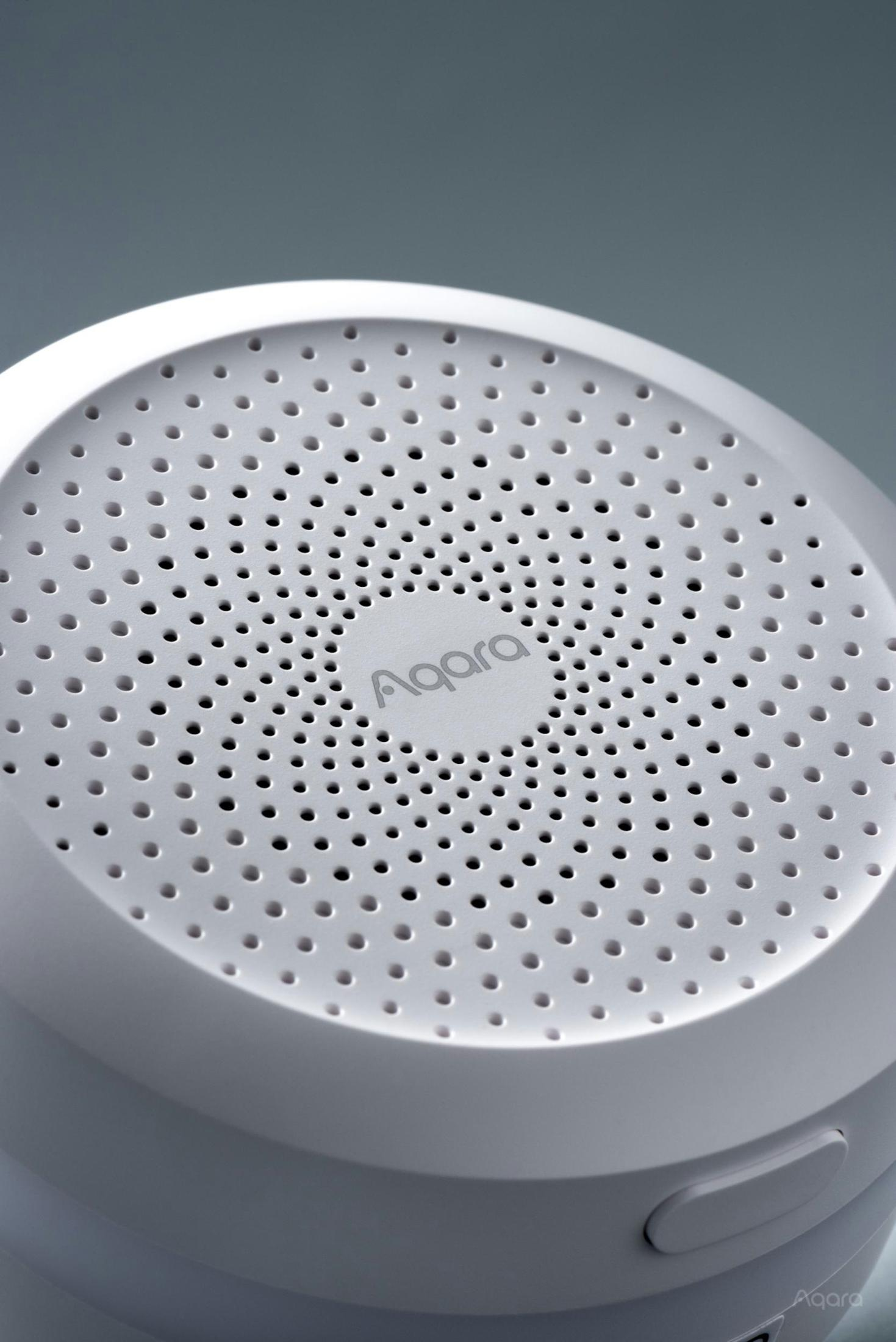 Nahaufnahme des weißen Aqara-Geräts. Perforierte Oberfläche mit dem Aqara-Logo in der Mitte. Grauer Hintergrund.