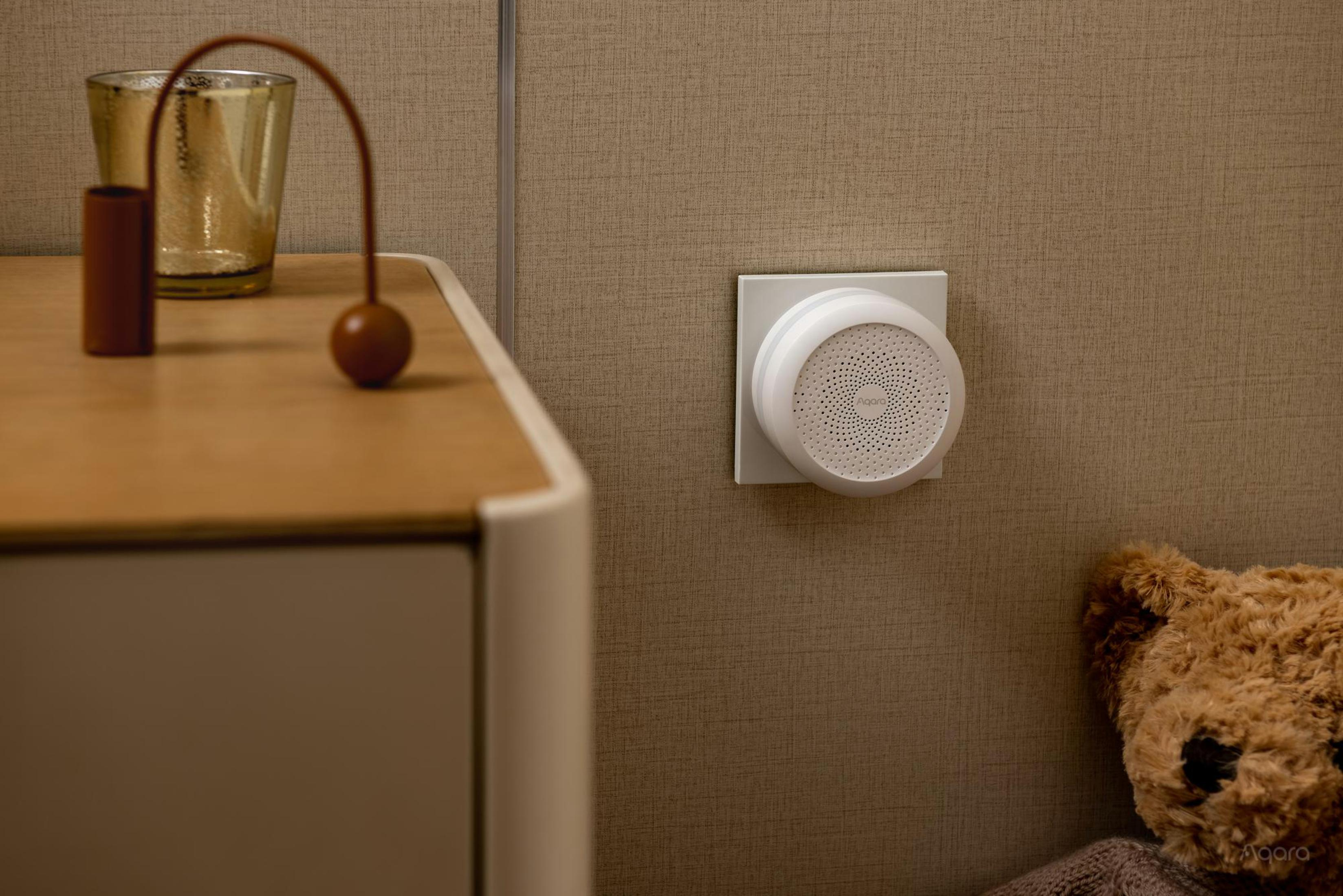 Wit Aqara-apparaat aan de muur. Daarnaast een houten nachtkastje met een kleine lamp en een teddybeer.
