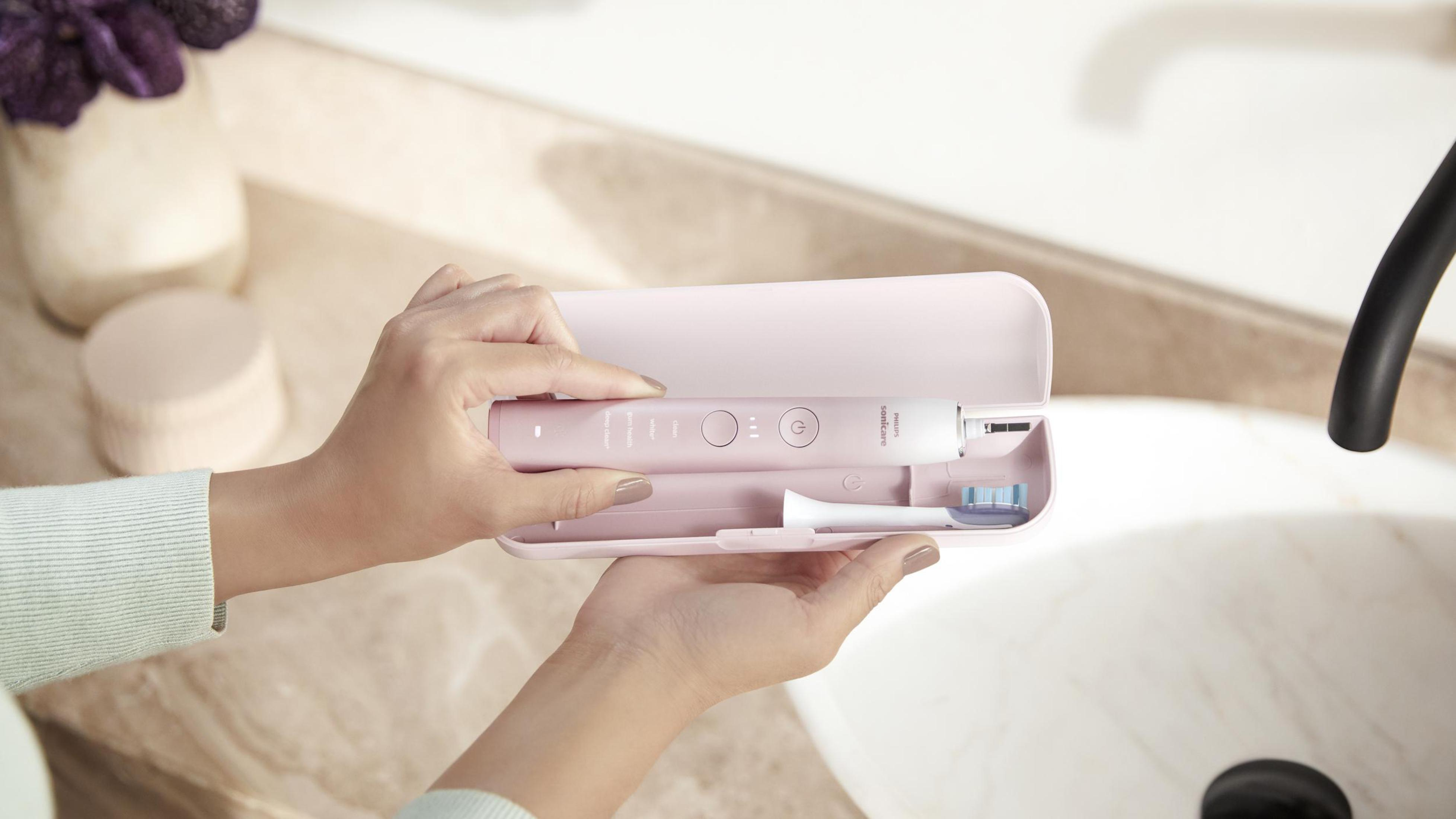 Person hält eine rosa elektrische Zahnbürste und ein Etui. Weißes Waschbecken und Spiegel im Hintergrund.