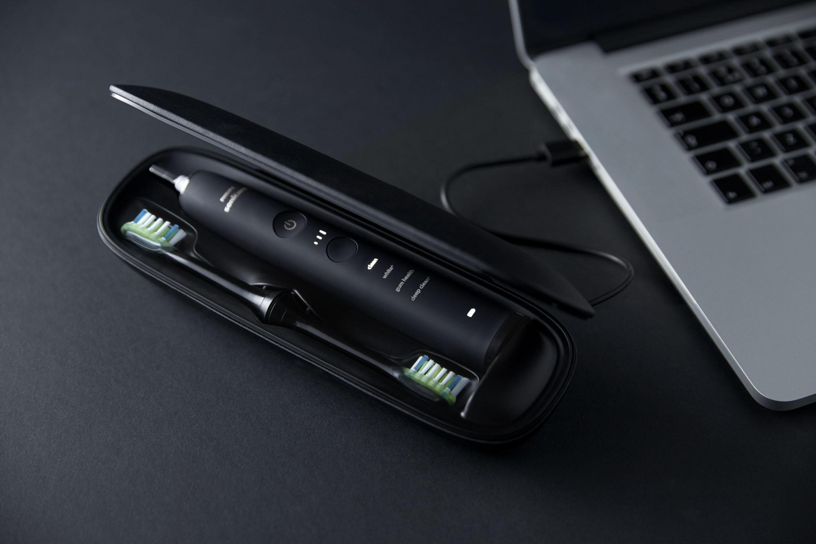 Black electric toothbrush in its case, open on a black surface next to a laptop, charging.