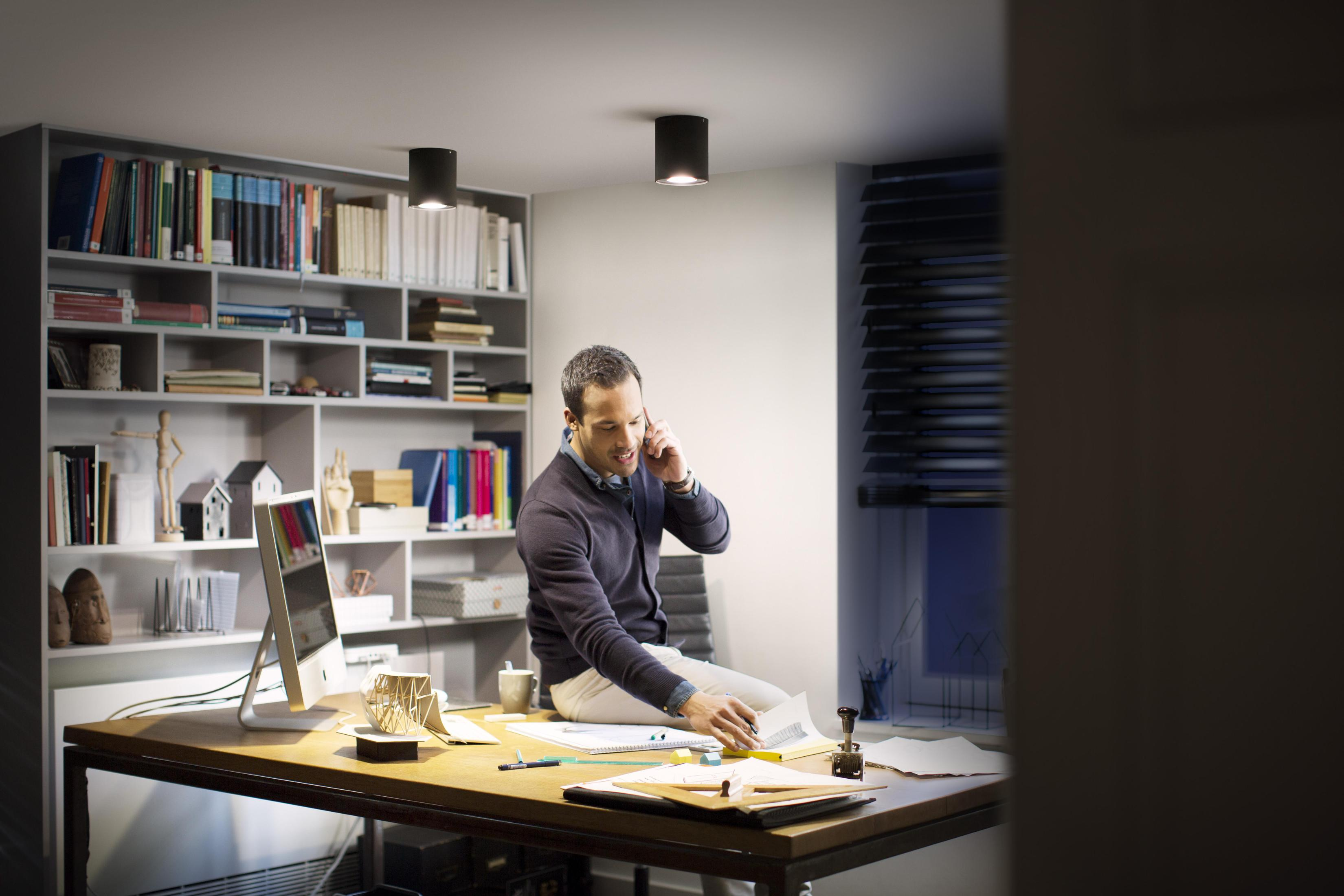 Mann sitzt in einem Büro auf einem Schreibtisch und telefoniert, Bücher in der Nähe.