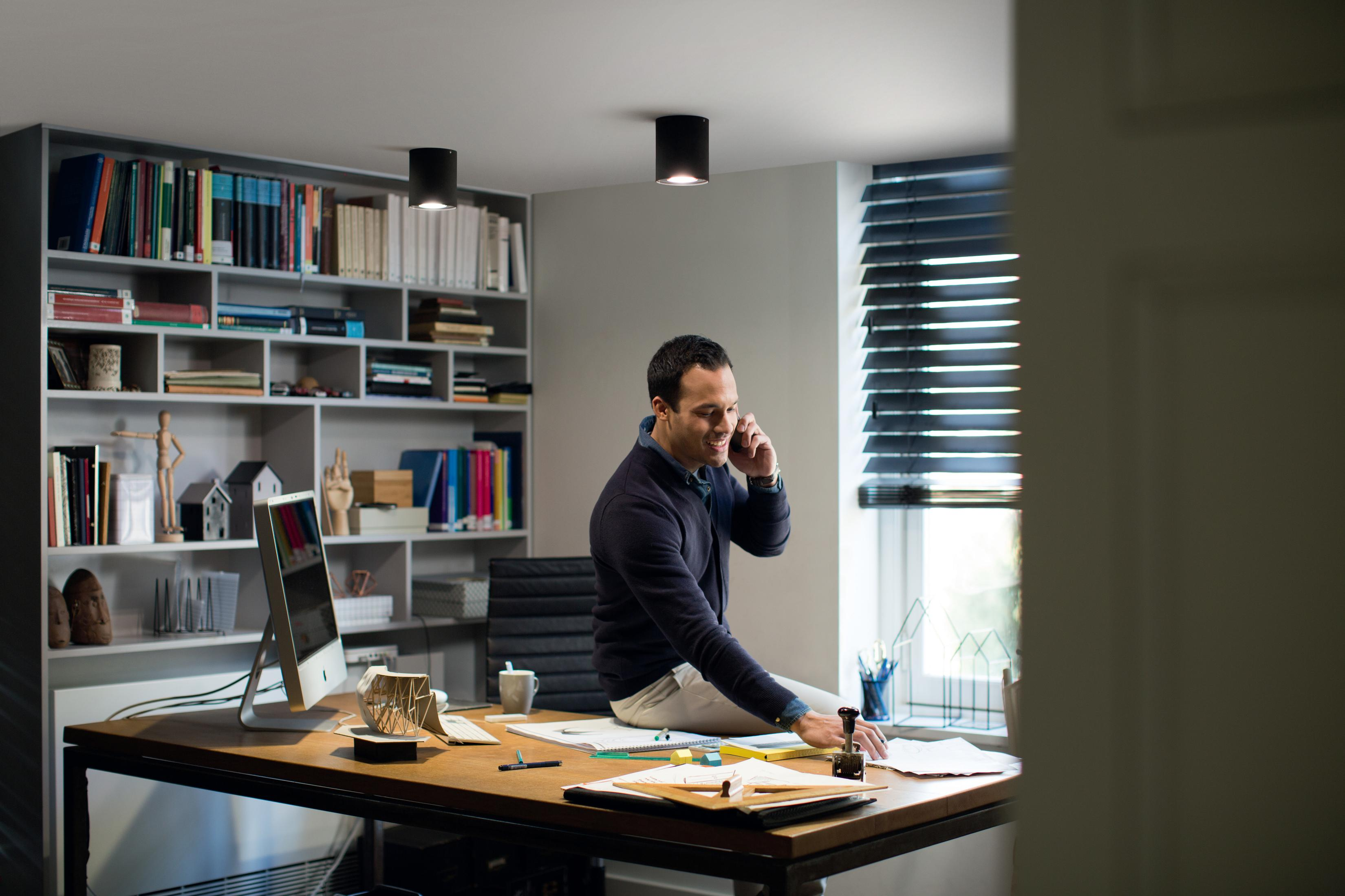 Ein Mann in einem blauen Pullover sitzt auf einem Schreibtisch und telefoniert in einem Büro.