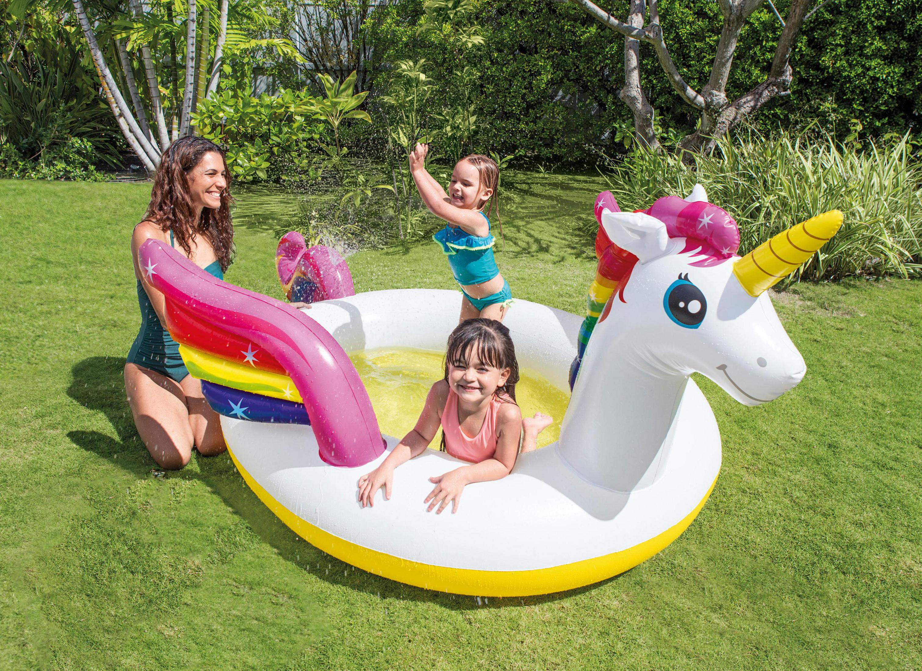Drei Kinder in einem Einhorn-Pool mit einer Frau. Grünes Gras und Bäume im Hintergrund.