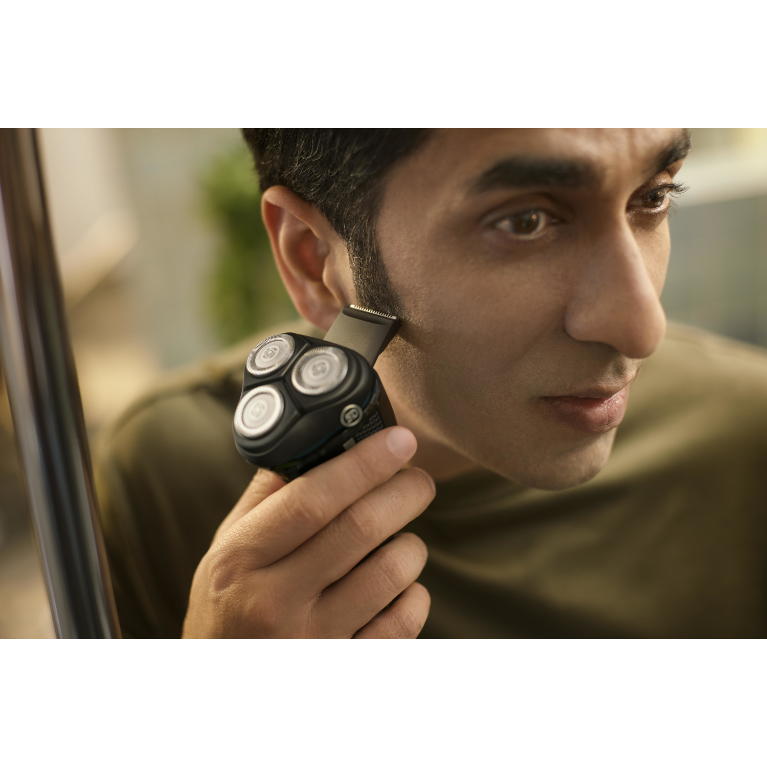 Mężczyzna goli bok twarzy czarną golarką elektryczną. Golarka ma trzy okrągłe głowice.
