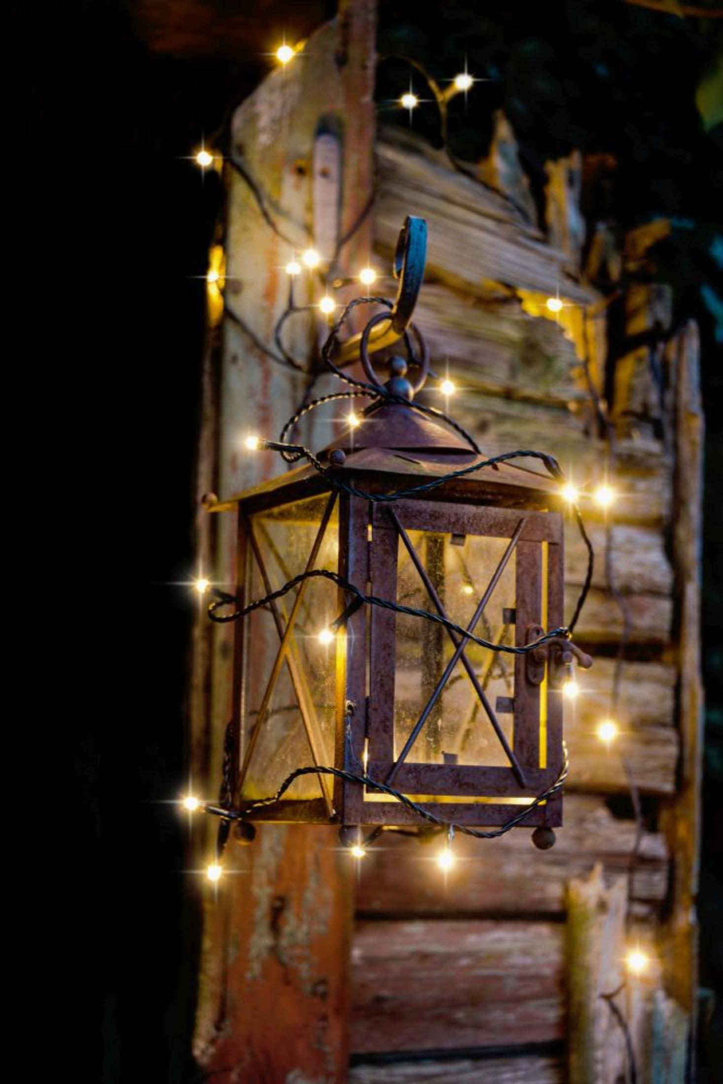Eine rustikale Laterne hängt an einem Holzpfosten, beleuchtet von Lichterketten, die einen warmen, einladenden Schein erzeugen.