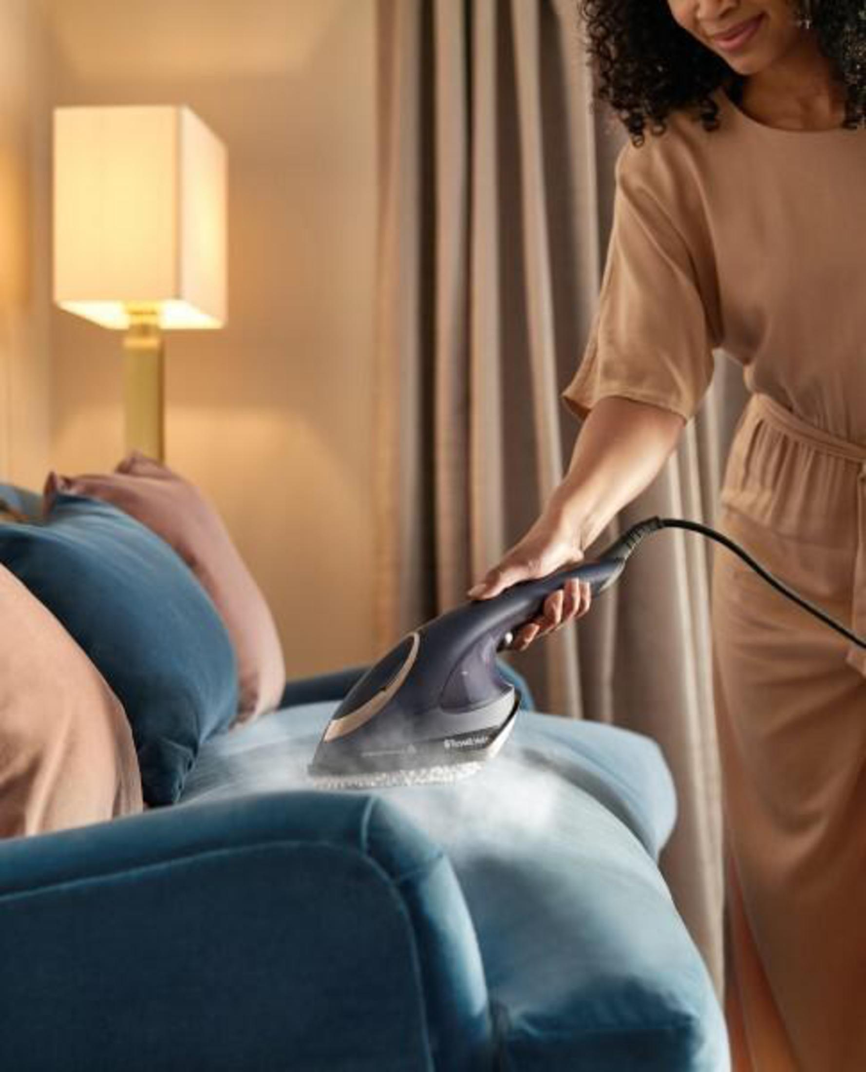 Kobieta używa parownicy ręcznej na niebieskiej sofie. Beżowa sukienka i lampa w tle.