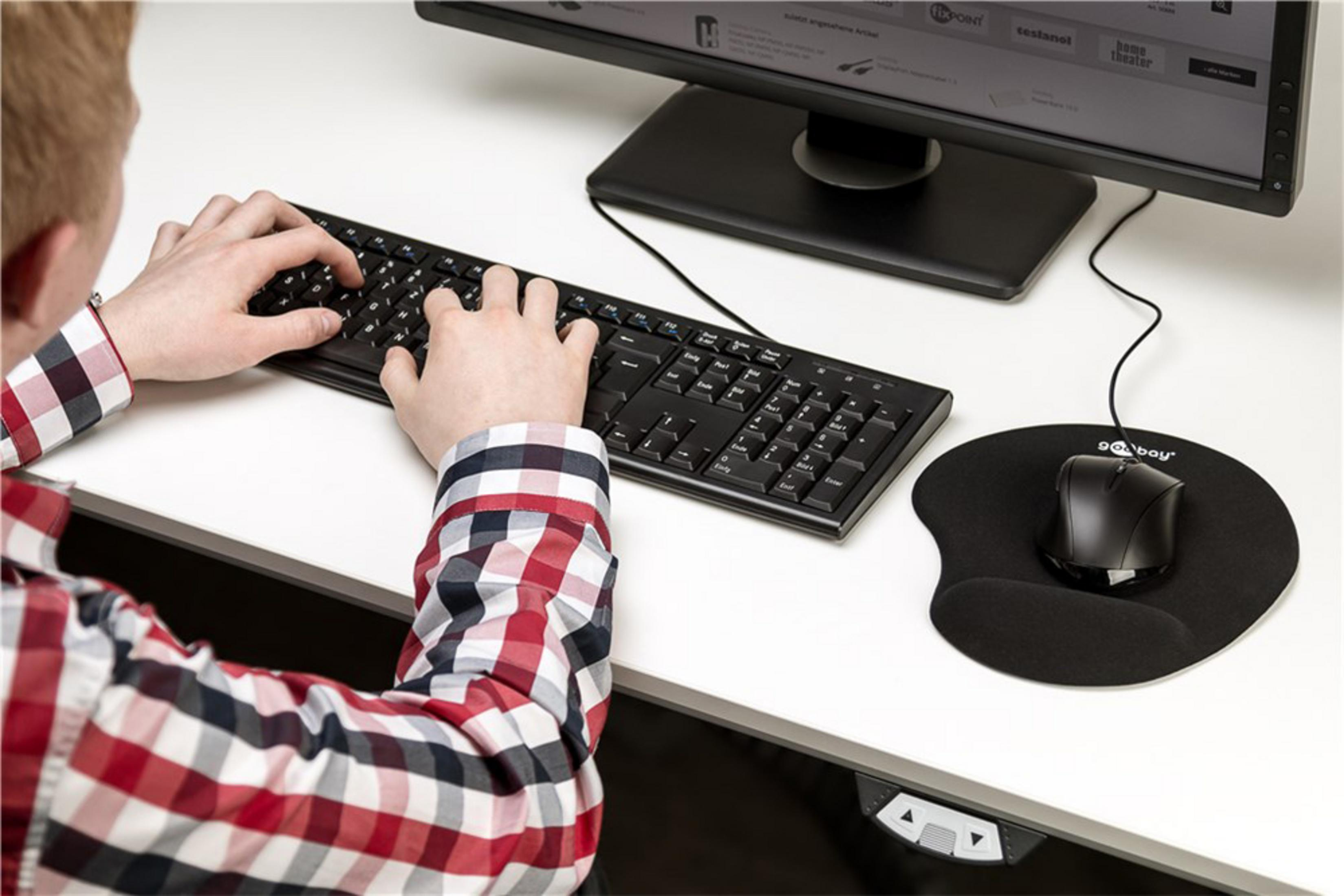 Eine Person benutzt eine schwarze Tastatur und Maus auf einem weißen Schreibtisch, mit einem Computermonitor.