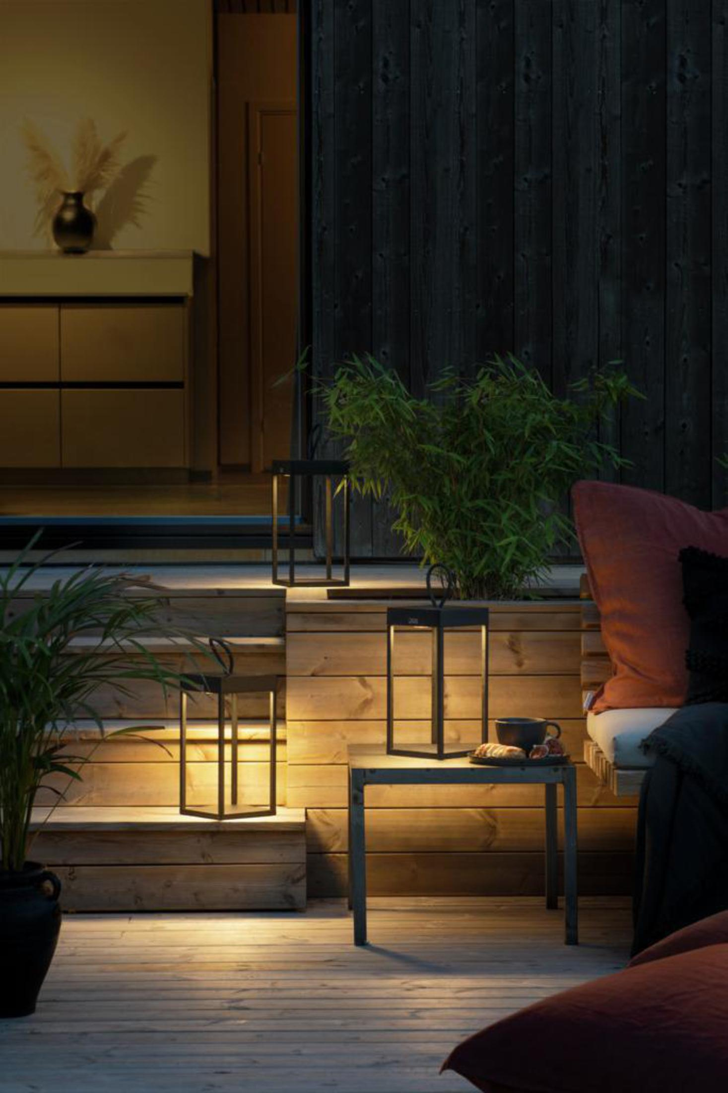 Außenszene bei Nacht mit beleuchteten Laternen und kleinem Tisch. Holz und Pflanzen.