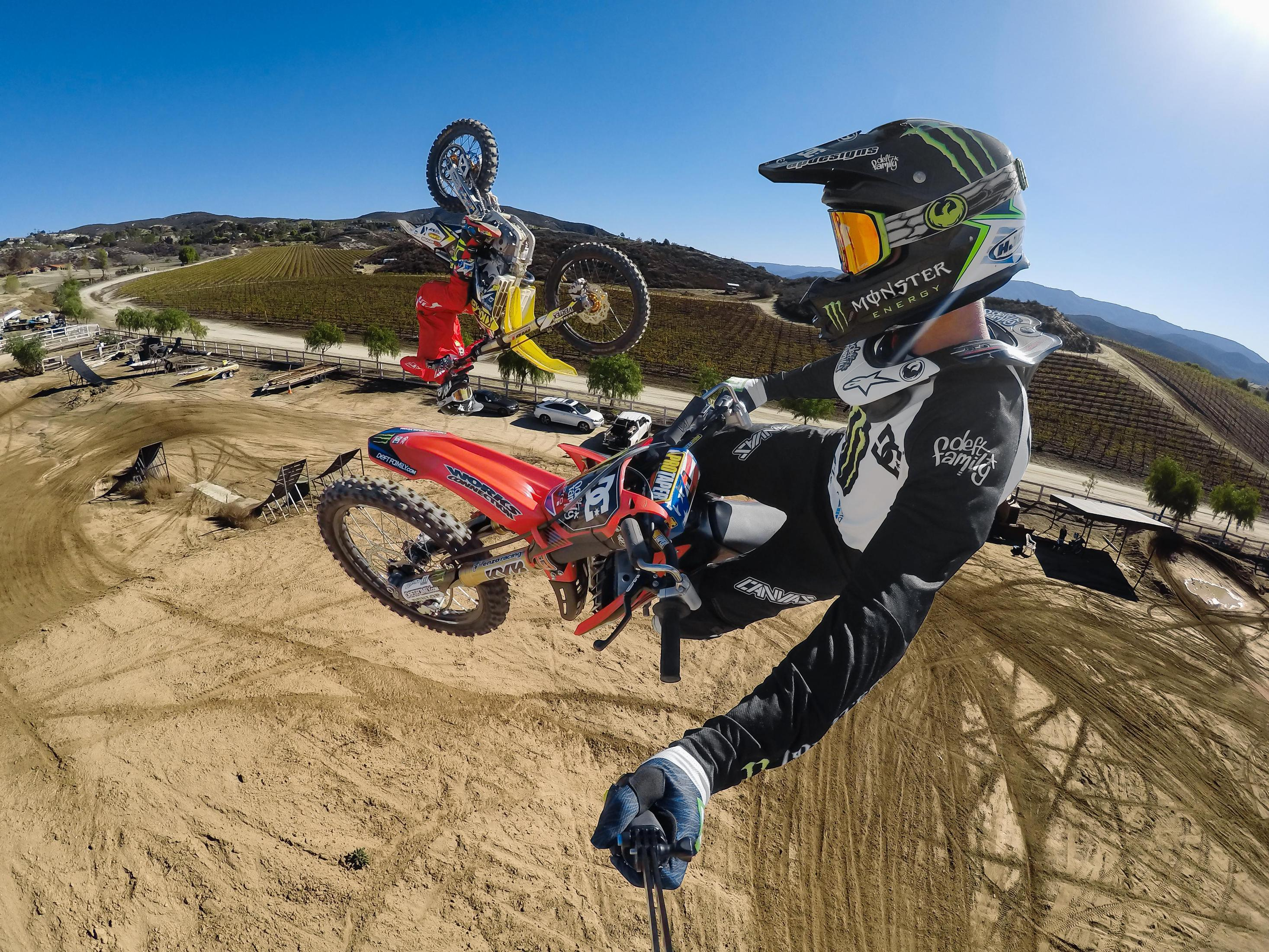 Piloto de motocross en el aire en una moto roja y blanca, otro piloto detrás, cielo azul y pista de tierra.