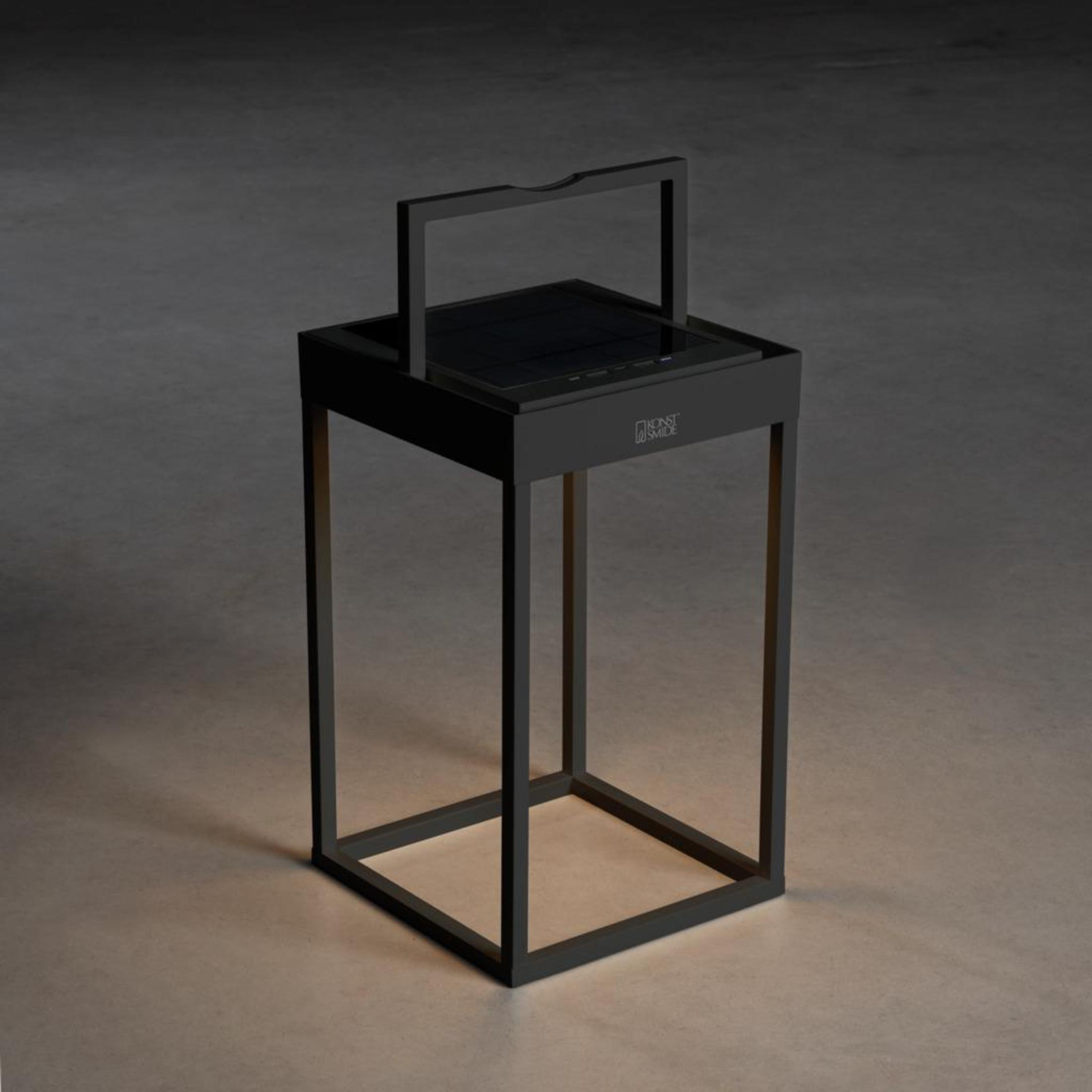 Schwarze Solar-Außenlaterne, rechteckige Form, mit Griff und Licht oben, auf dunkler Oberfläche.