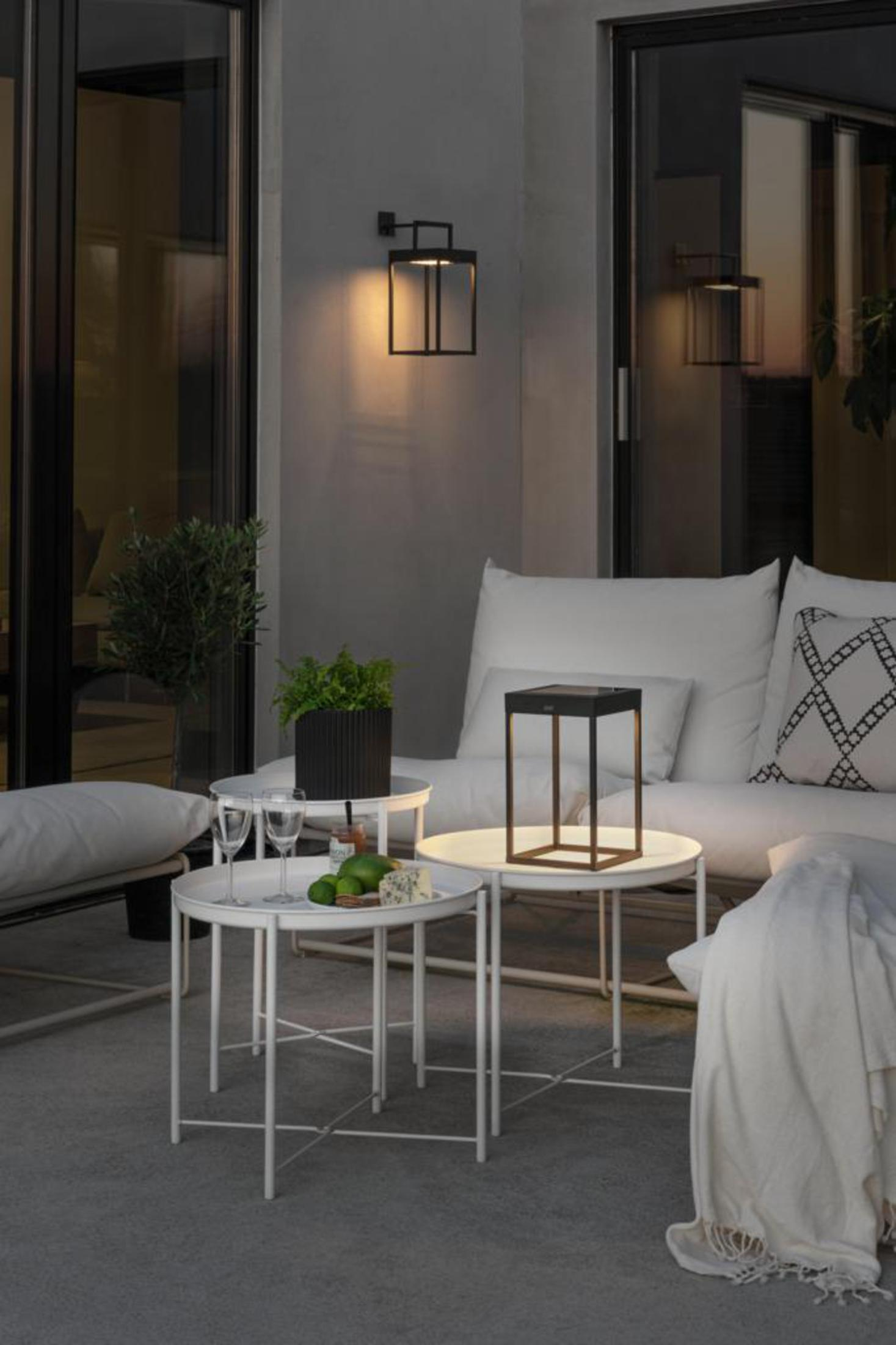 Außenszene in der Dämmerung mit weißem Sofa, Beistelltischen, einer Wandleuchte und einer Tischlampe.