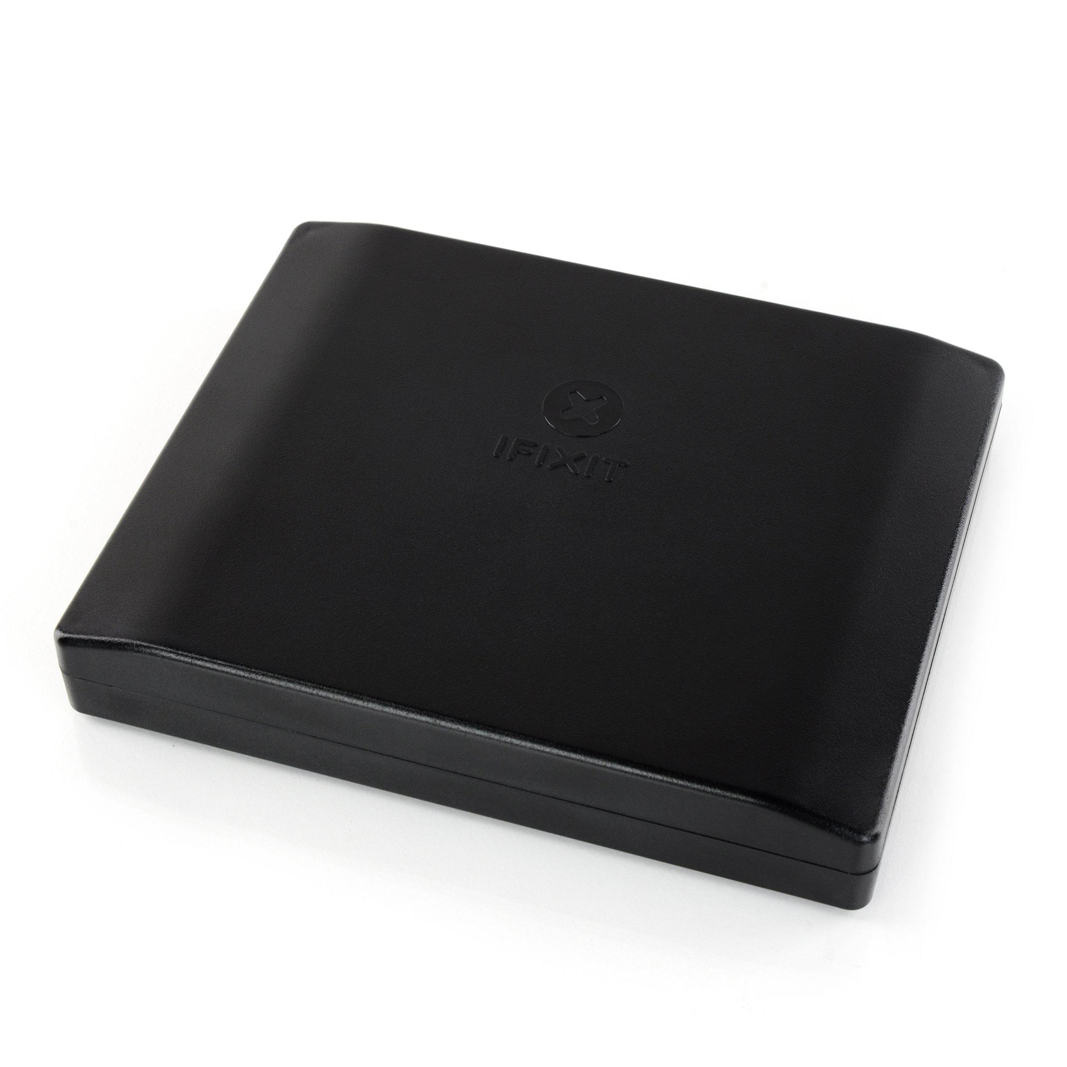 Eine schwarze iFixit-Box ist geschlossen. Das iFixit-Logo ist sichtbar. Sie liegt auf einer weißen Oberfläche. Die Box ist leicht angewinkelt.
