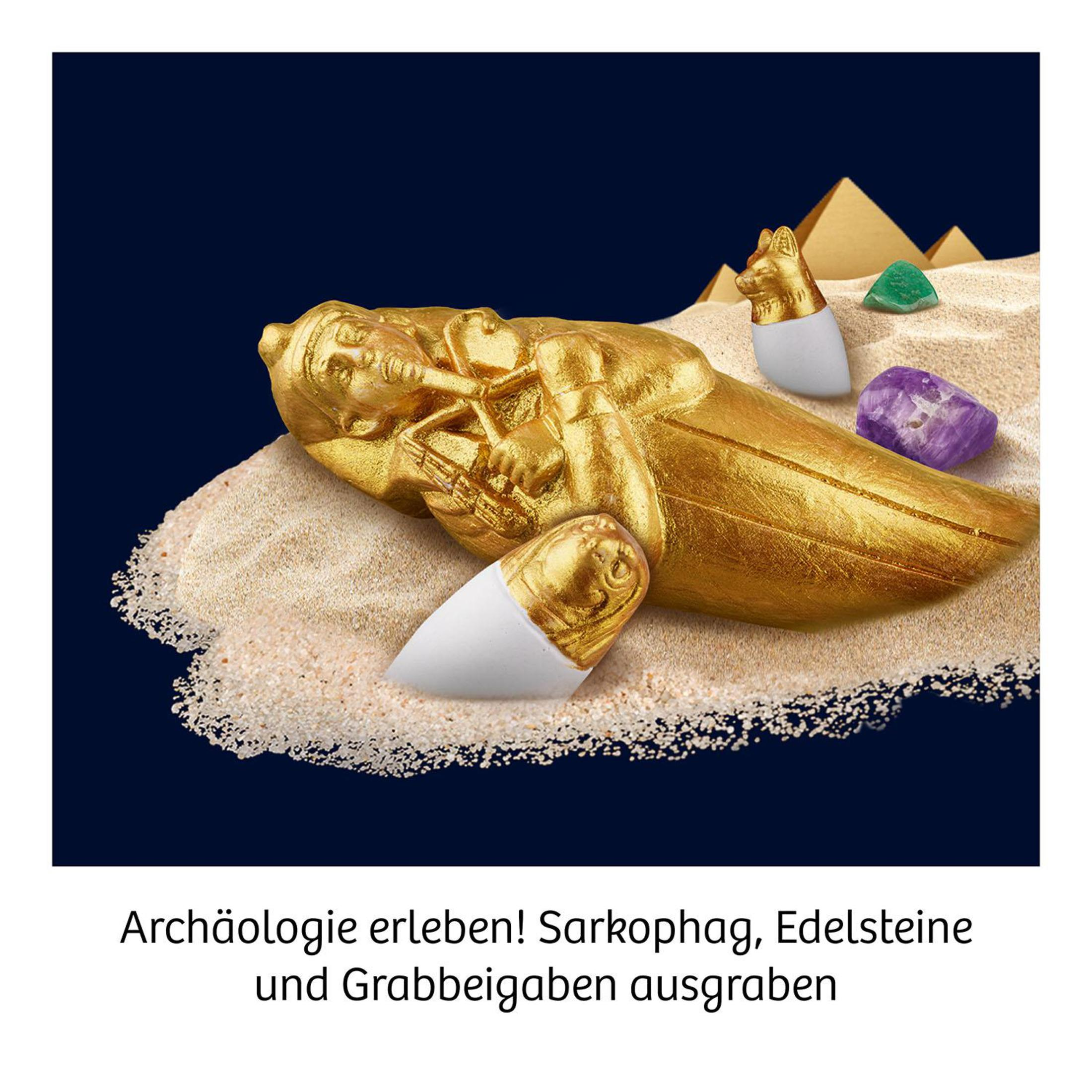Ein goldener Sarkophag mit Edelsteinen und ägyptischen Artefakten auf Sand, mit Text darunter.
