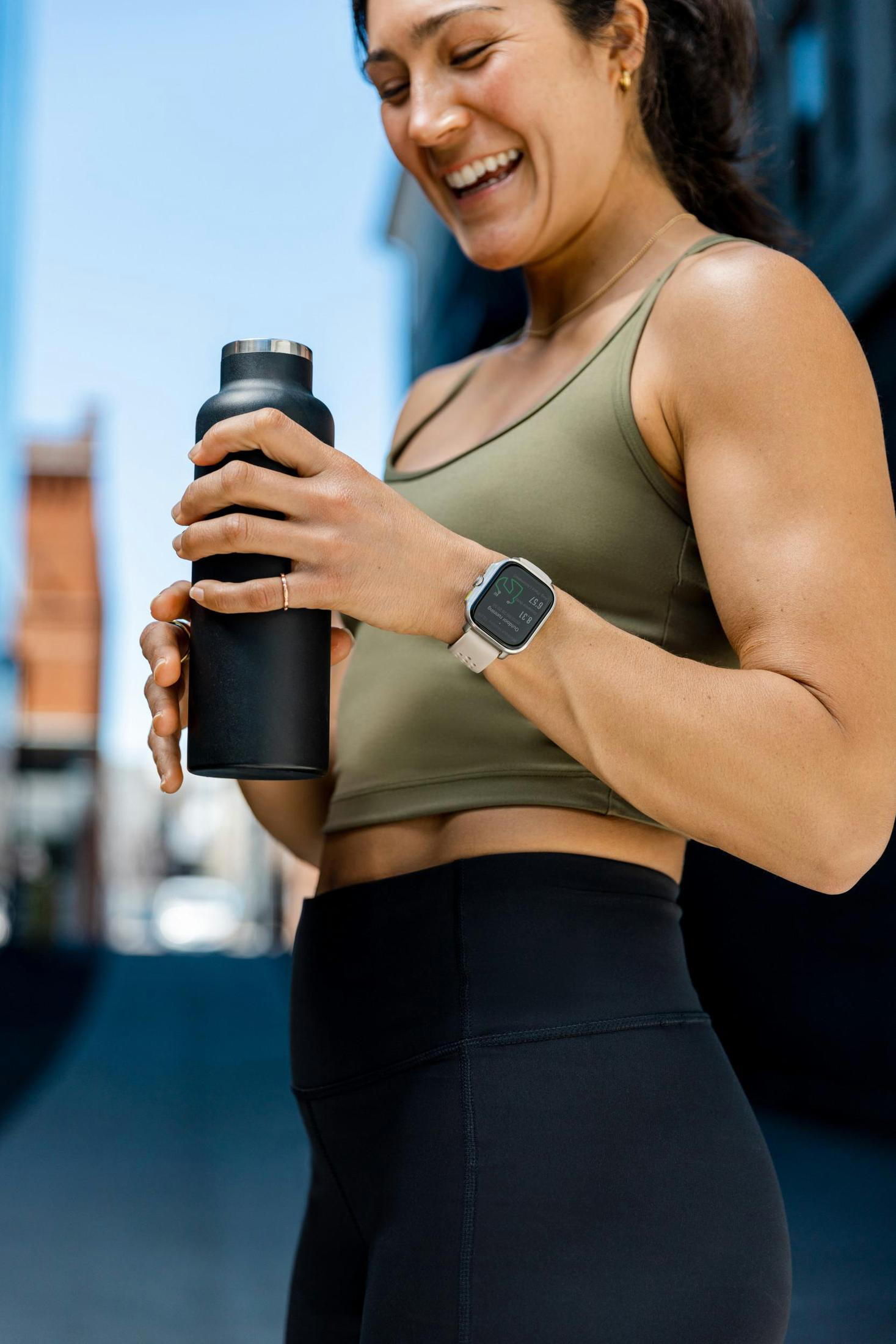 Kobieta w stroju sportowym uśmiecha się, trzyma czarną butelkę, smartwatch, tło miejskie.