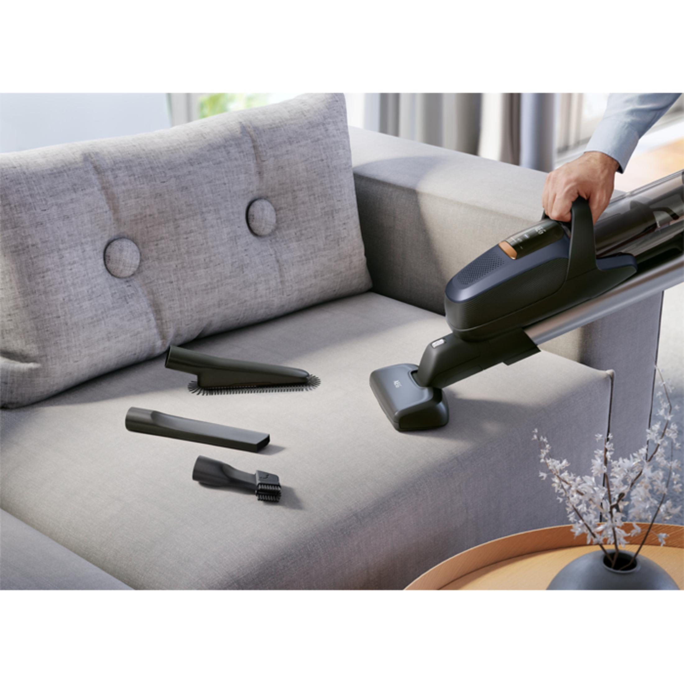 Odkurzacz czyszczący sofę. Czarny odkurzacz z nakładkami na szarej sofie. Akcesoria obok. Białe tło.