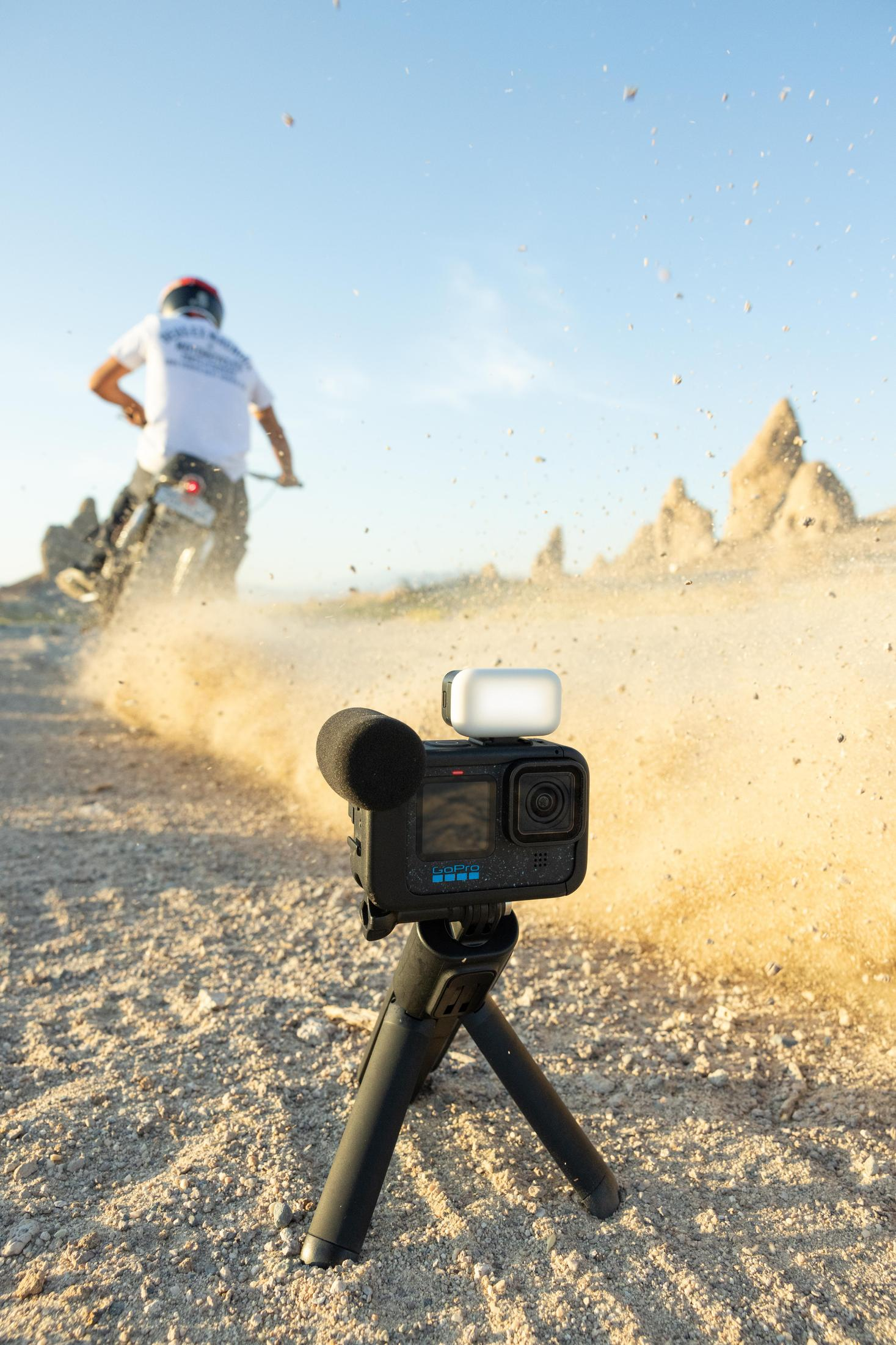 Kamera GoPro na statywie, nagrywająca motocyklistę na zakurzonej trasie z błękitnym niebem.